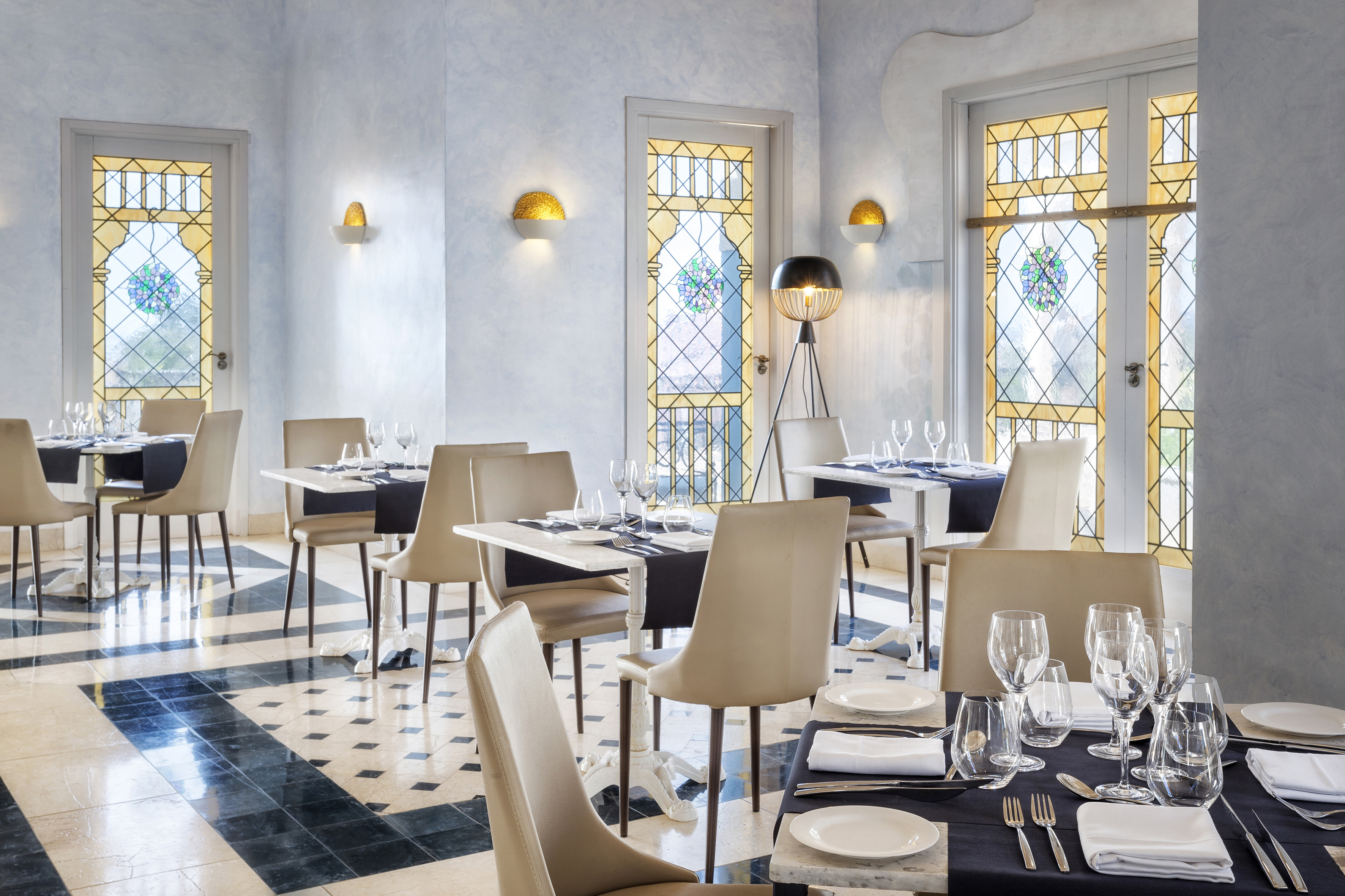 Restaurant dining room with set tables, light chairs, patterned floor, and stained glass.