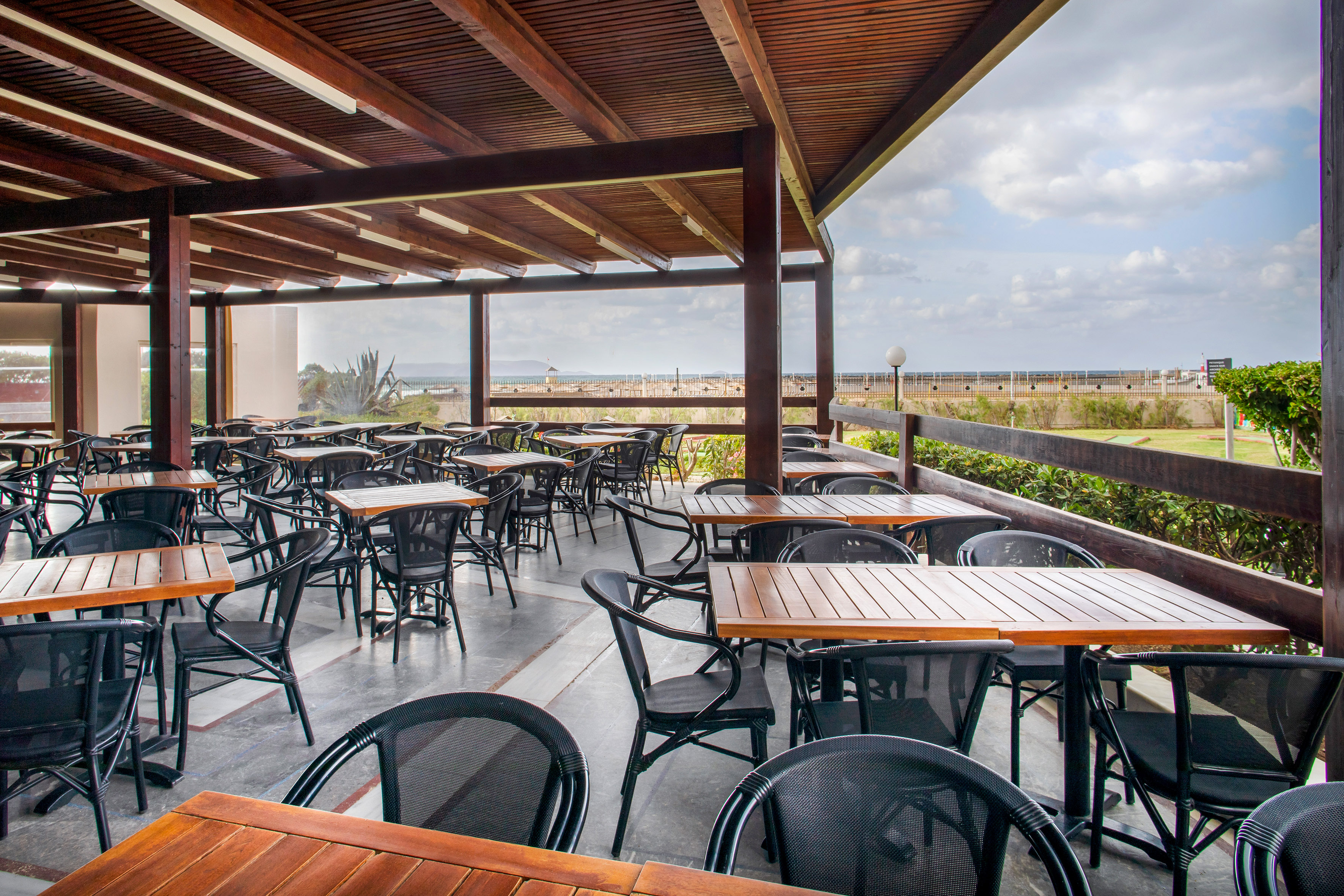 Outdoor restaurant terrace with wooden tables, black chairs, and sea view.
