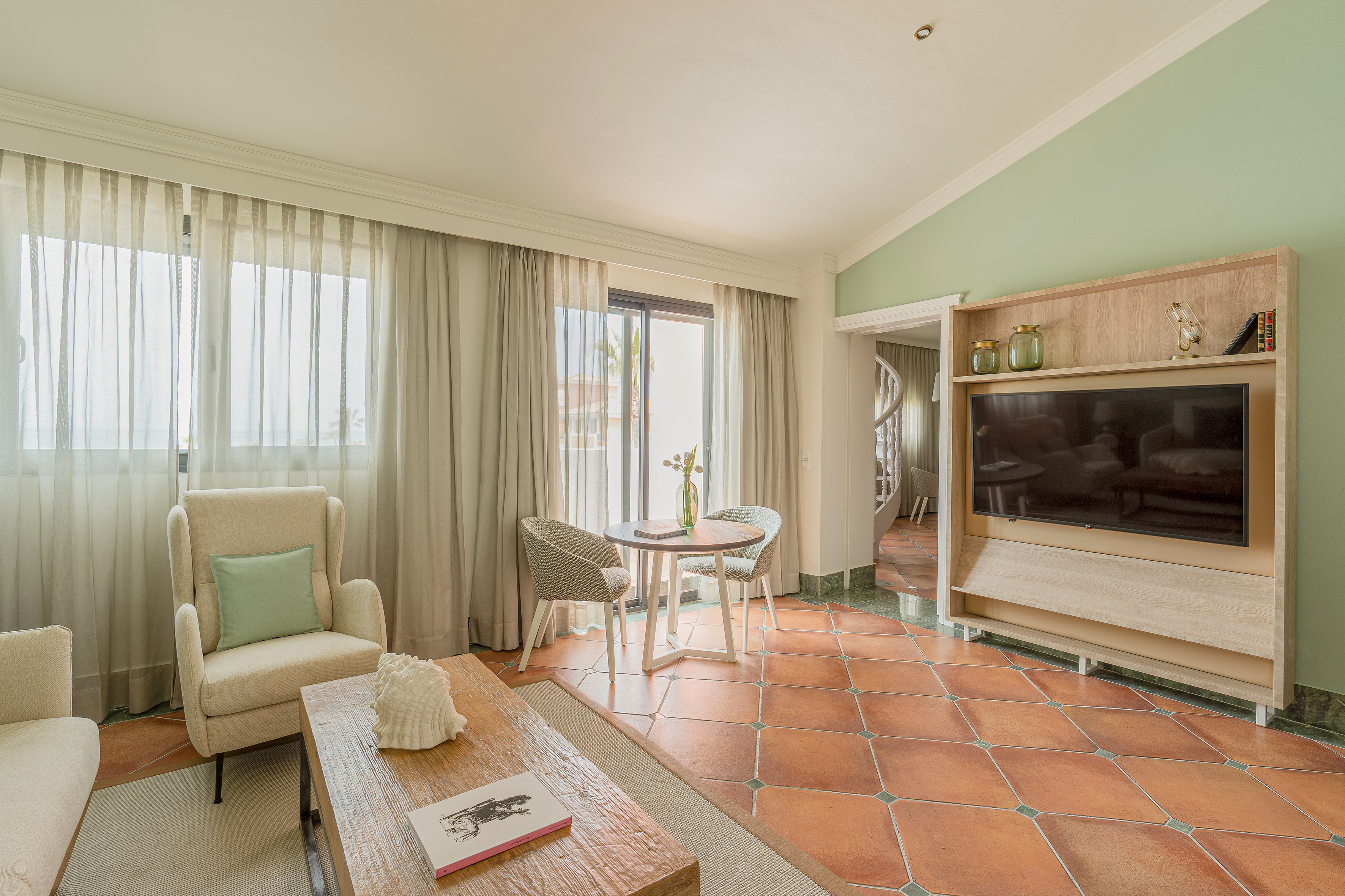 Modern hotel suite living room with terracotta floor, green wall, and balcony.