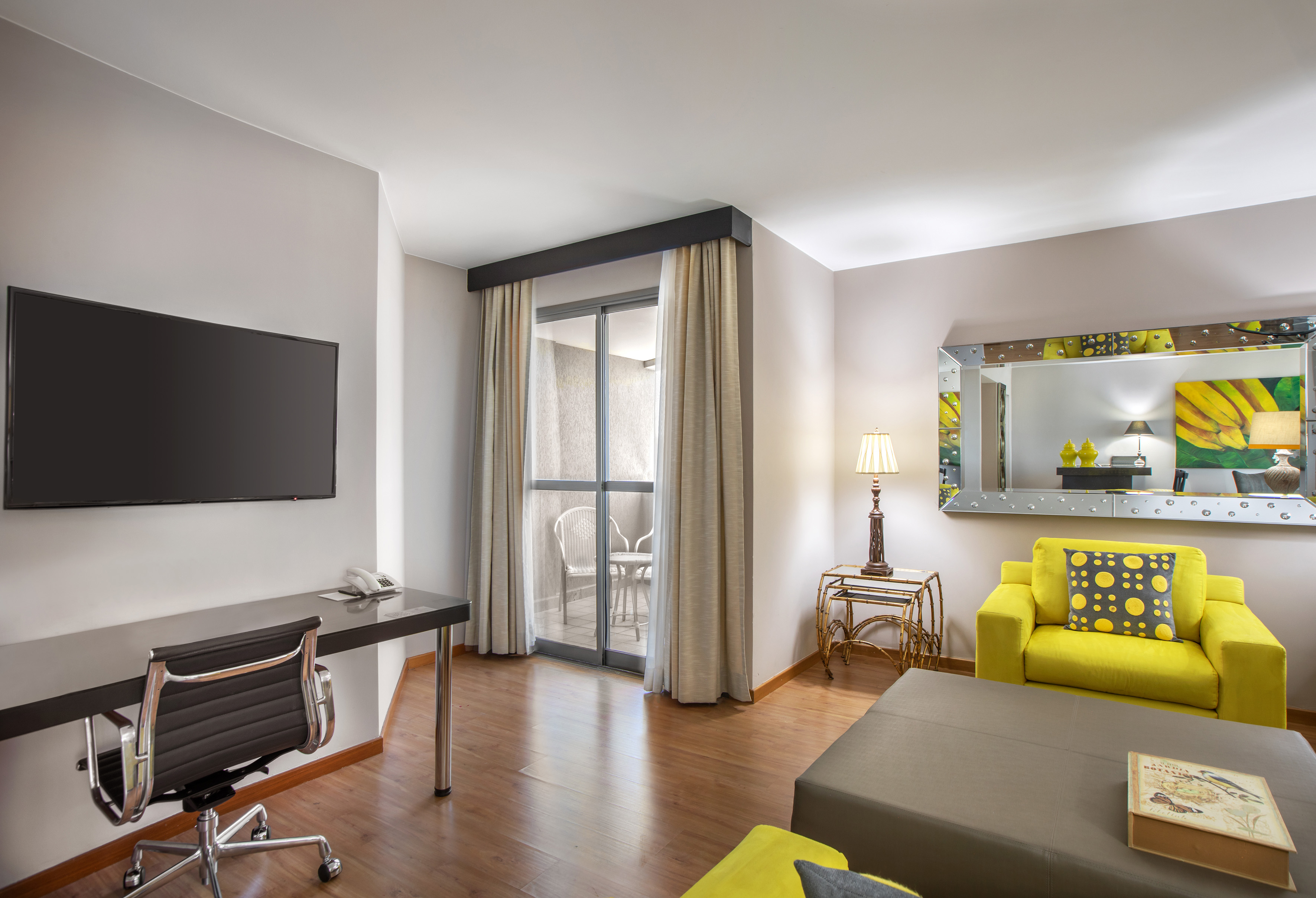 Modern hotel suite with balcony, yellow armchair, TV, and desk.