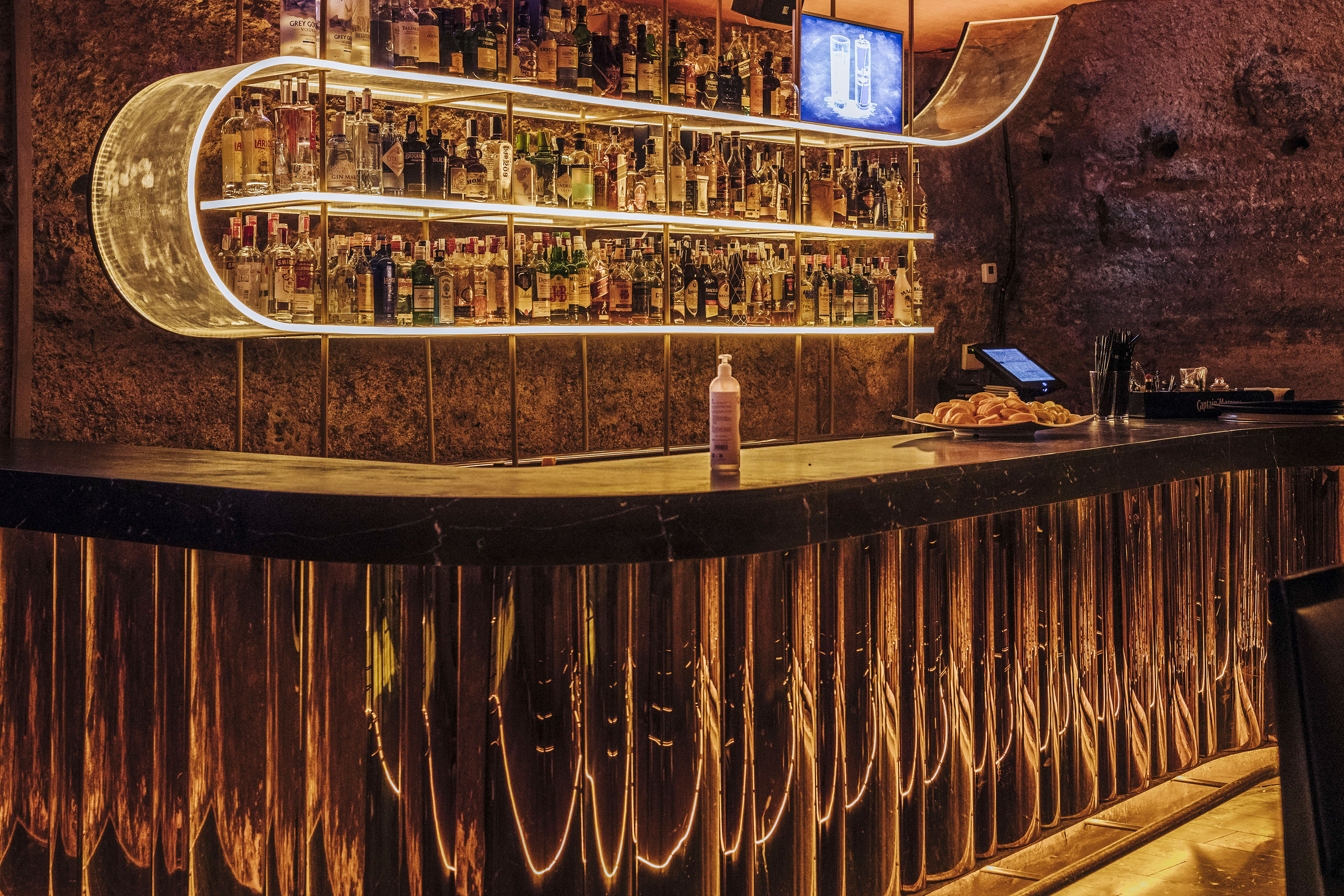 Illuminated modern bar with backlit liquor shelves, textured stone wall, and glowing counter.