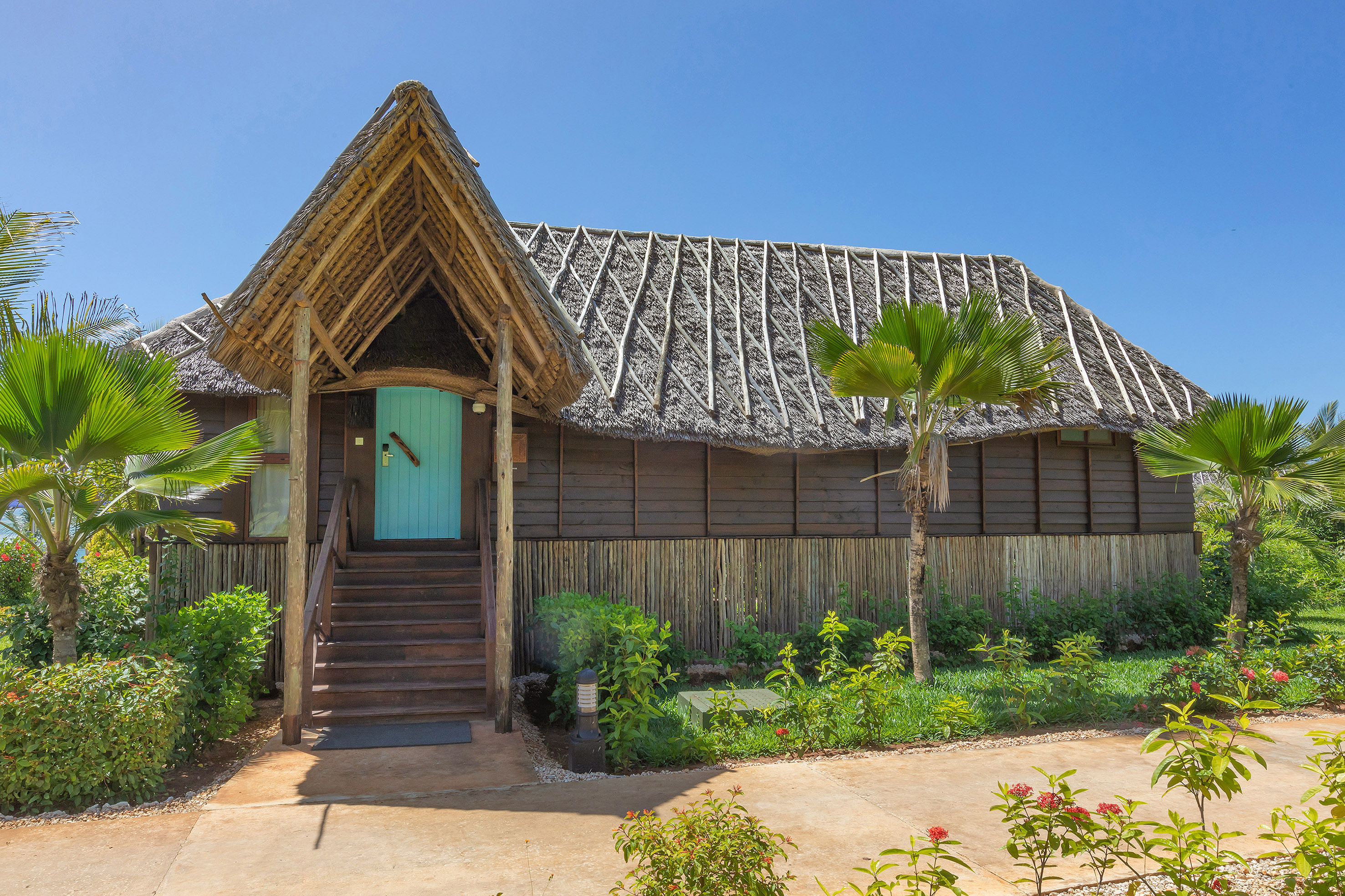 Wooden tropical bungalow with thatched roof, blue door, and palm trees.