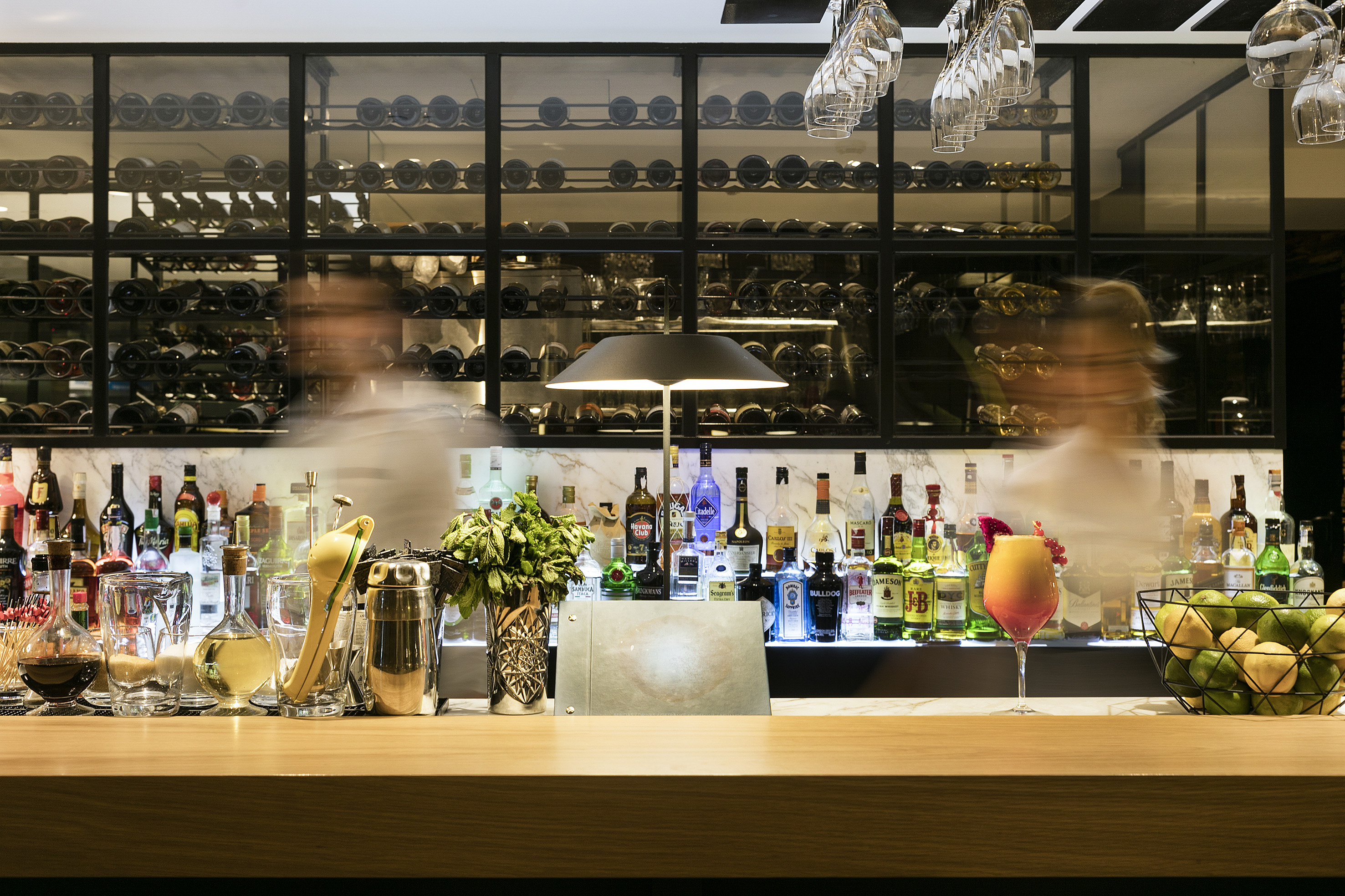 Bar counter with liquor bottles, wine display, and cocktail; blurred bartenders.