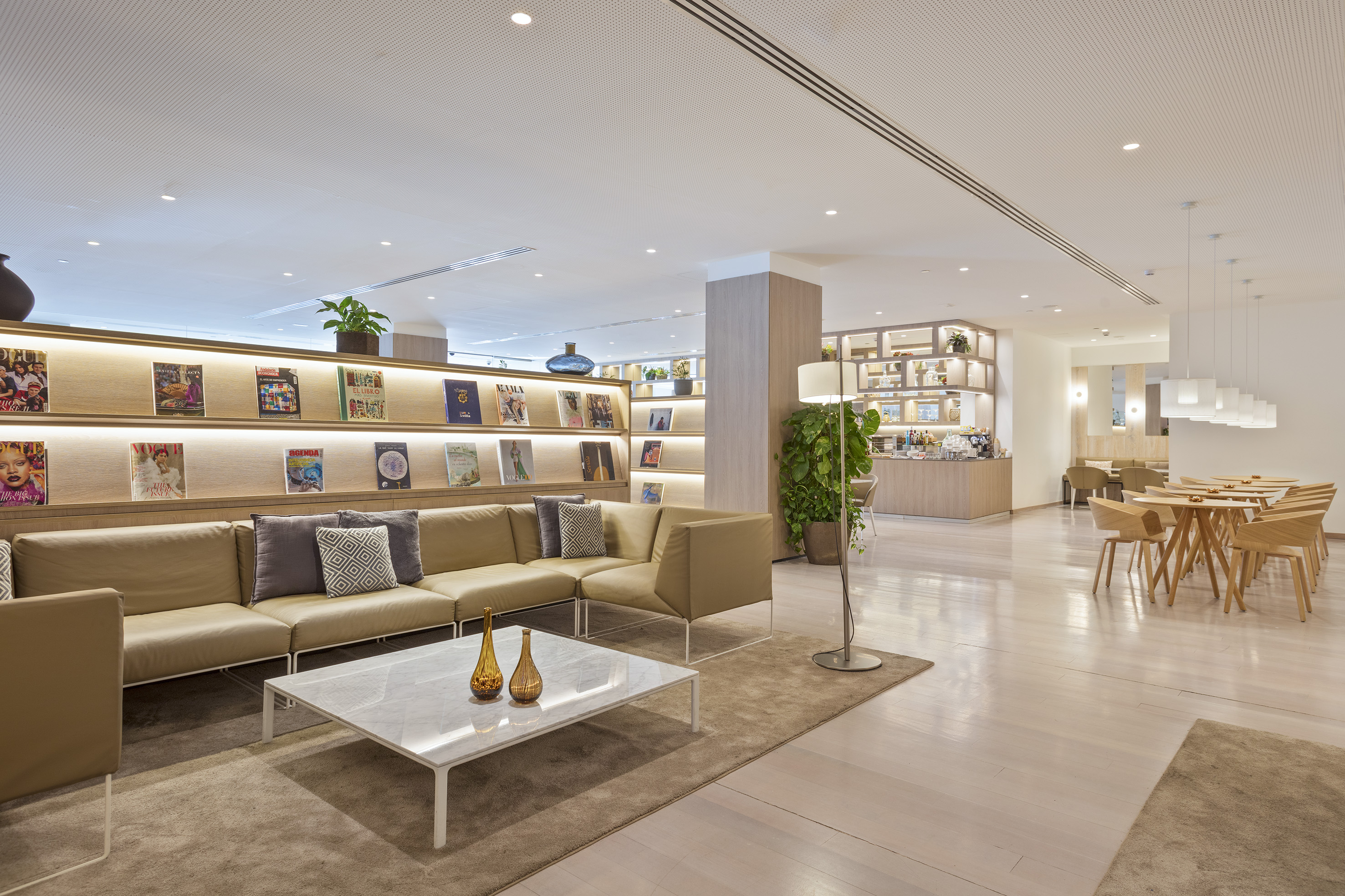 Modern hotel lobby lounge with tan sofas, magazine shelves, and dining area.