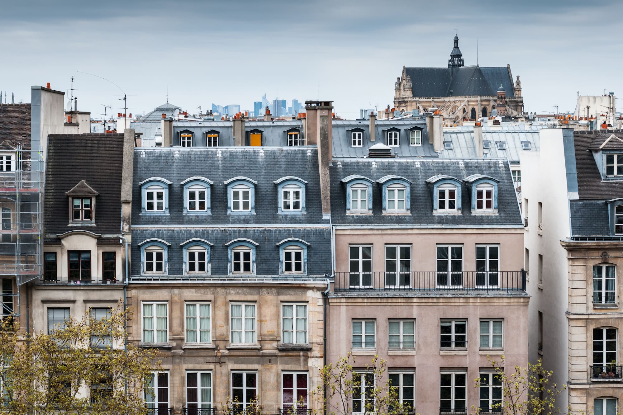 Edificios parisinos con tejados de pizarra, buhardillas y torre de iglesia al fondo.