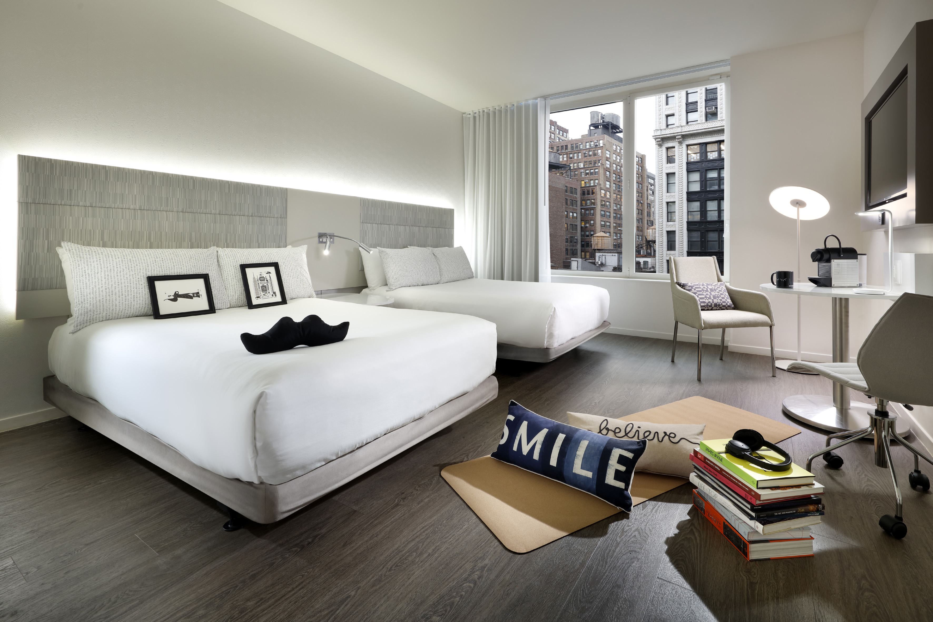 Modern hotel room with two beds, city view, desk, and contemporary decor.