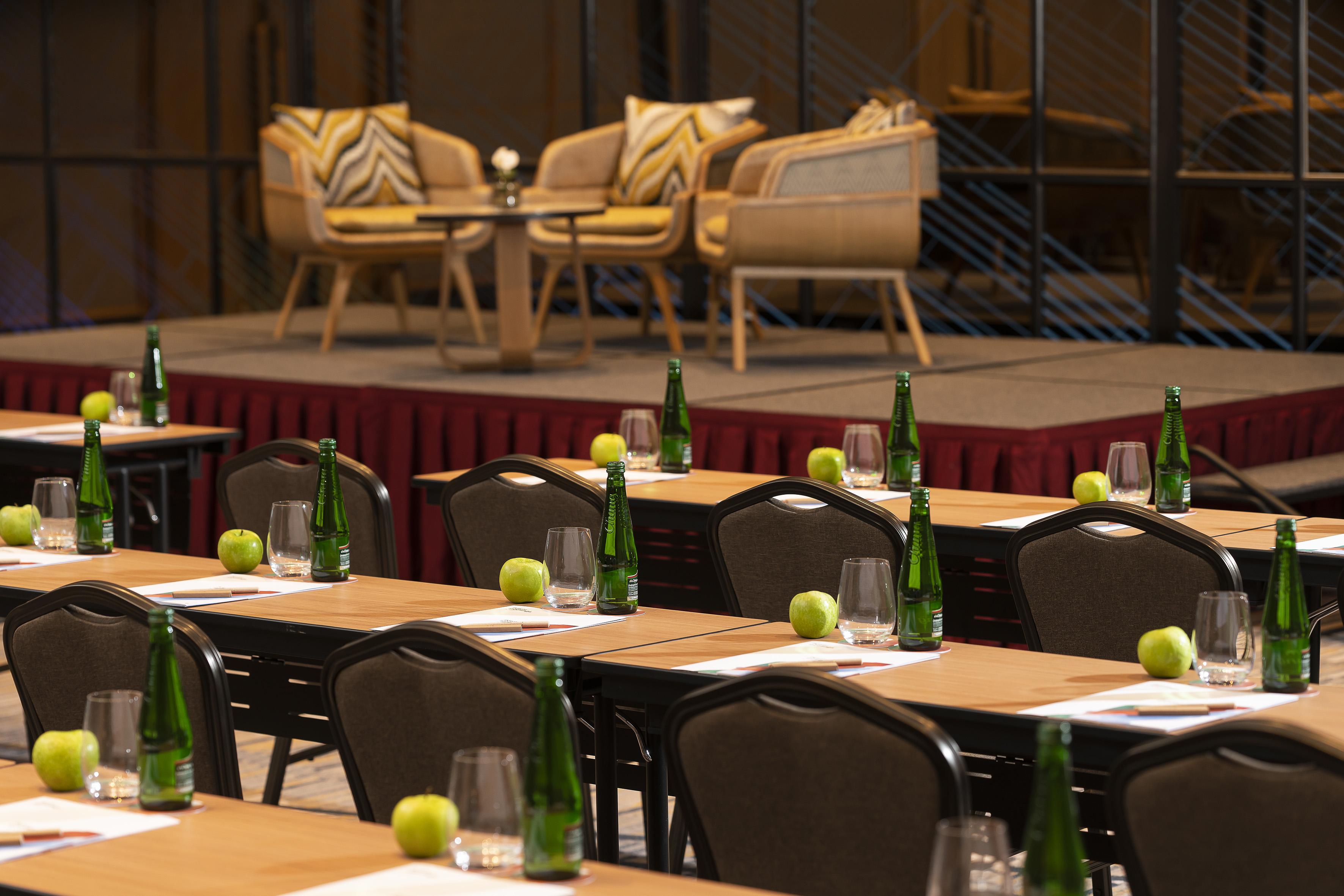 a room with tables and chairs and a stage with chairs and a stage with chairs and a table with green bottles and apples