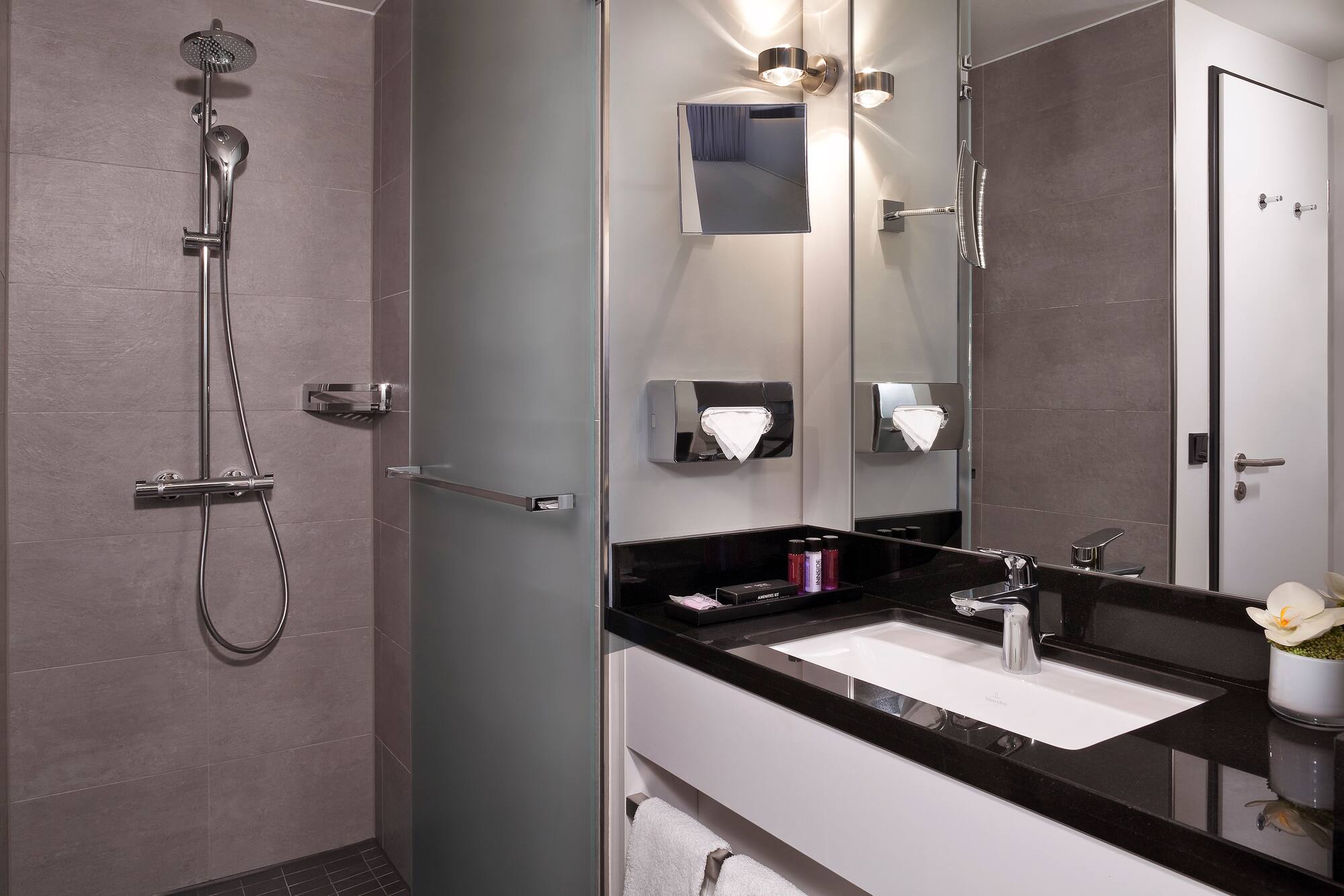 Modern hotel bathroom: grey tiled walk-in shower, black vanity, white sink.