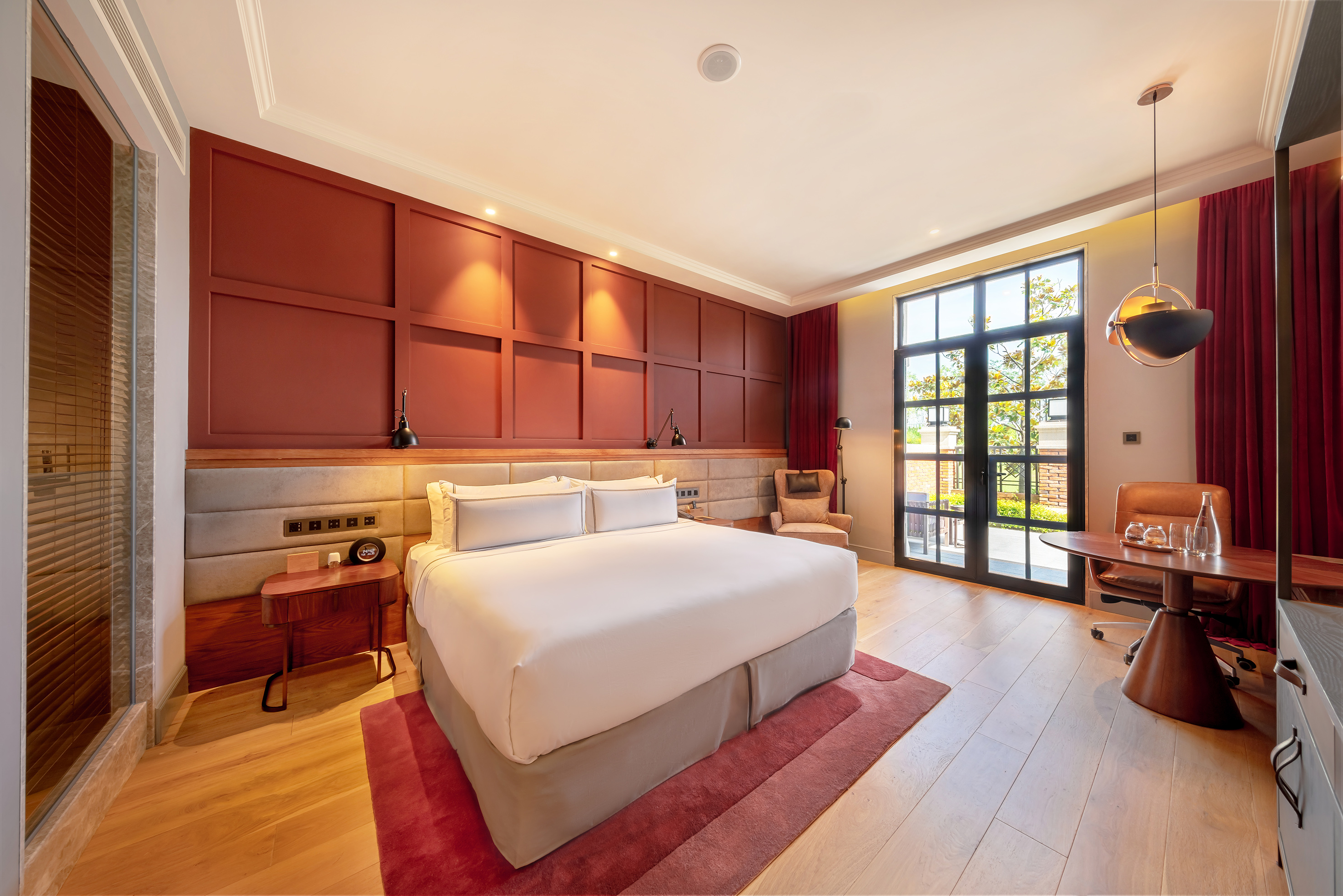 Elegant hotel suite with king bed, red wall, desk, and natural light.