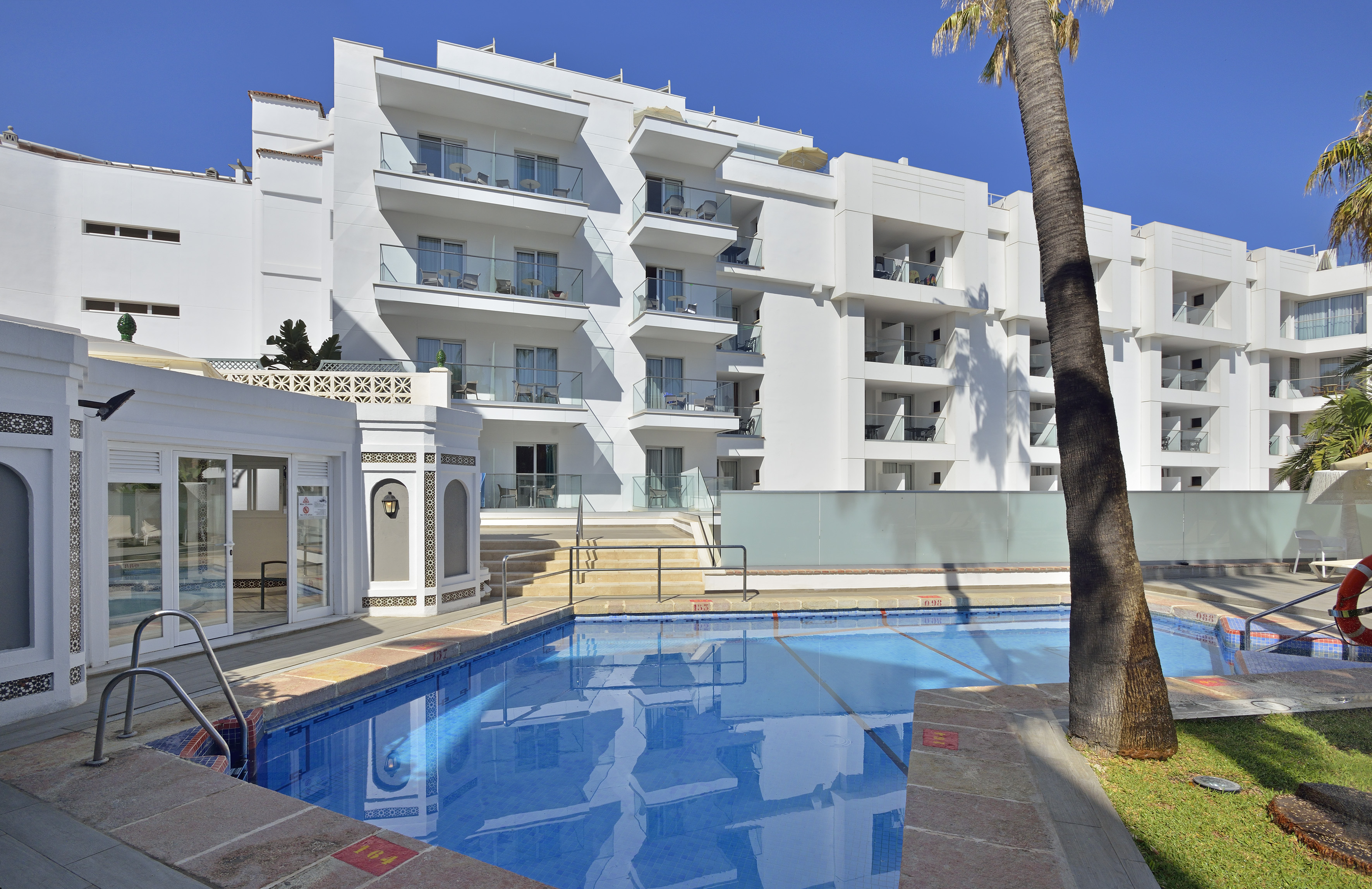 White hotel building with balconies, large outdoor swimming pool, and palm tree.