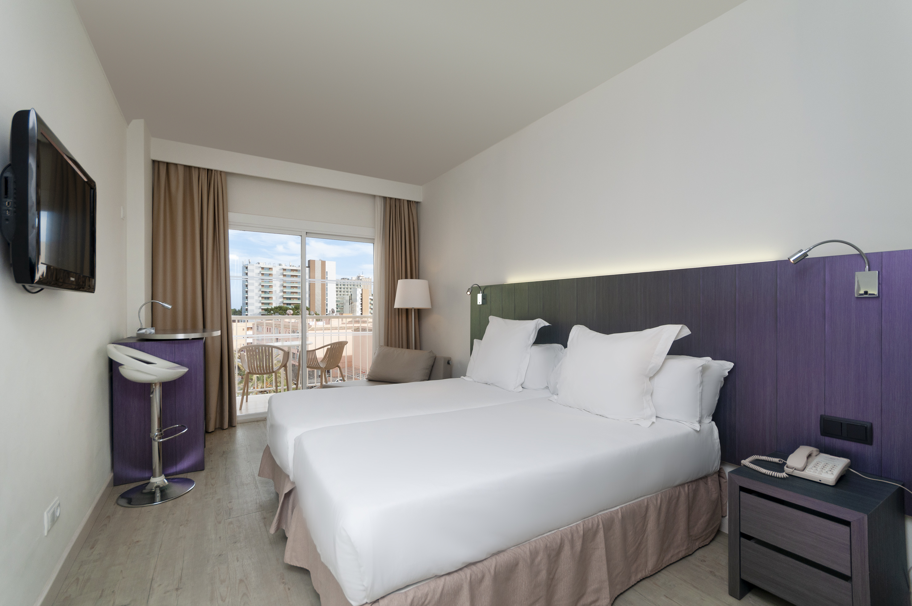 Modern hotel room with twin beds, balcony, wall TV, and purple headboard.