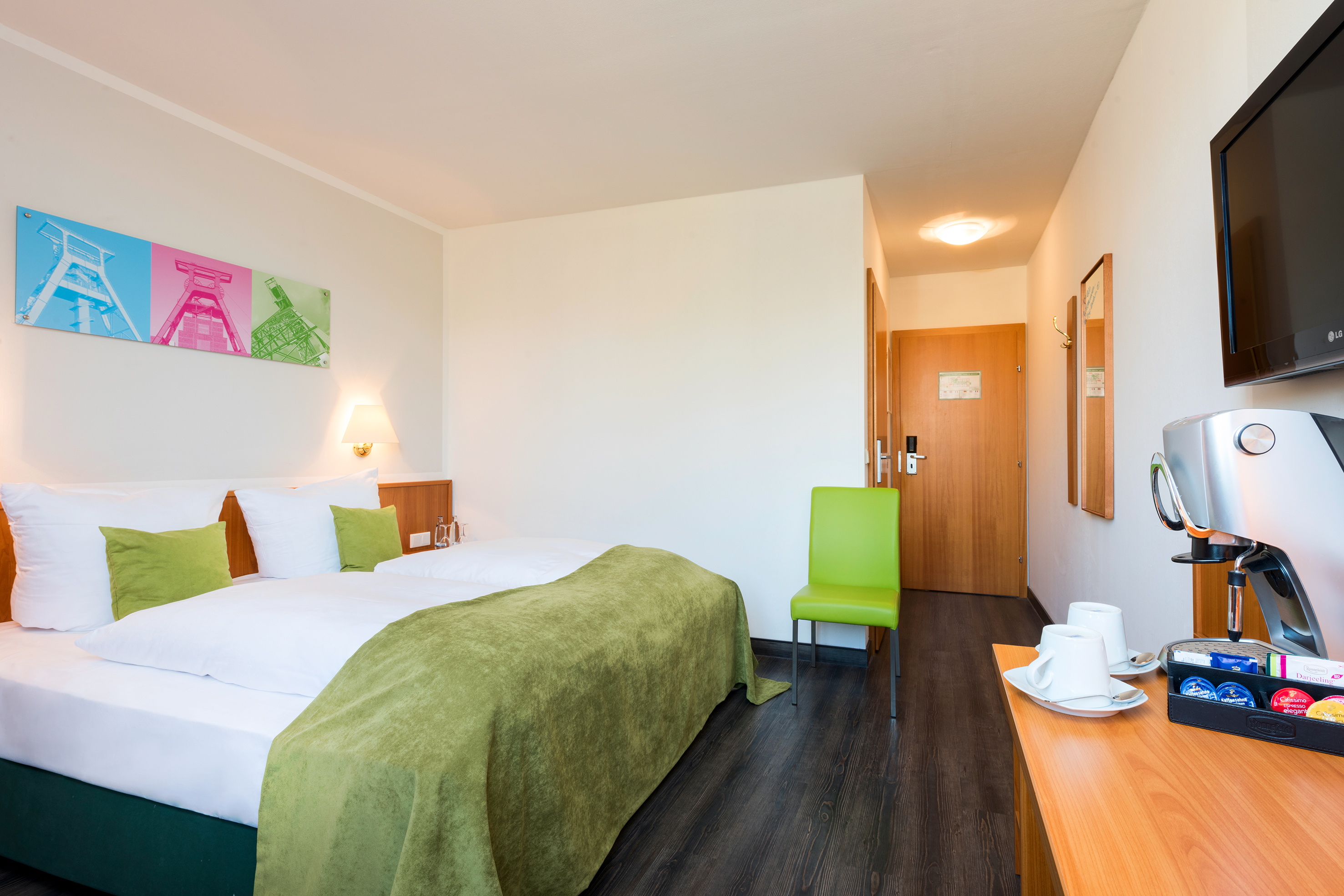 Bright hotel room with double bed, green bedding, coffee station, mining art.