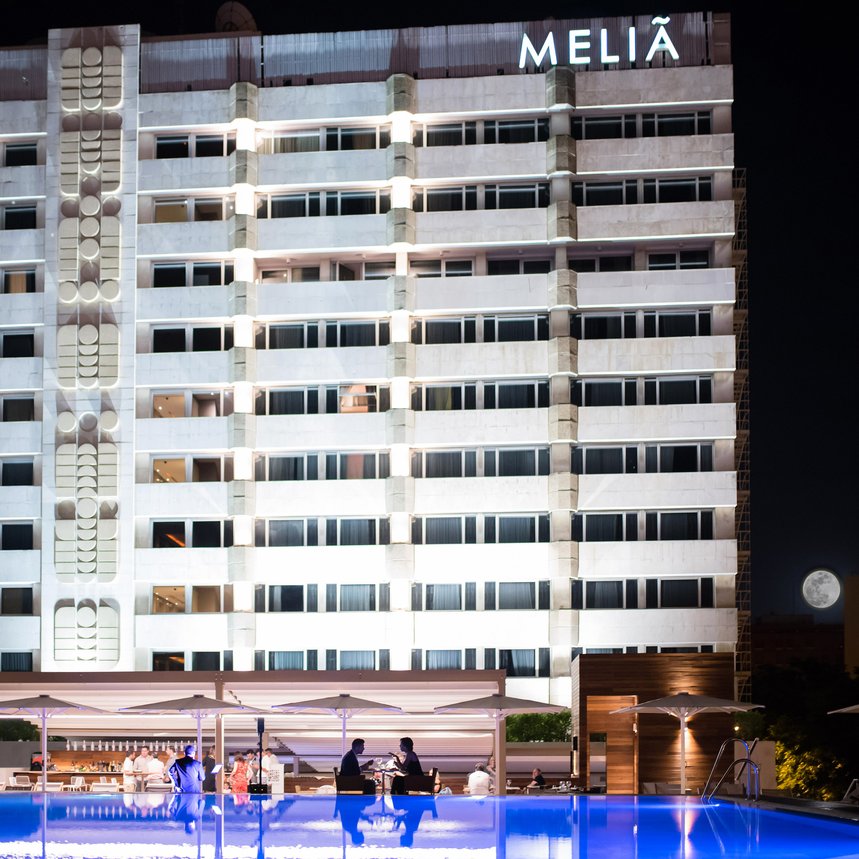 Melia hotel lit at night, with vibrant pool, outdoor dining, and full moon.