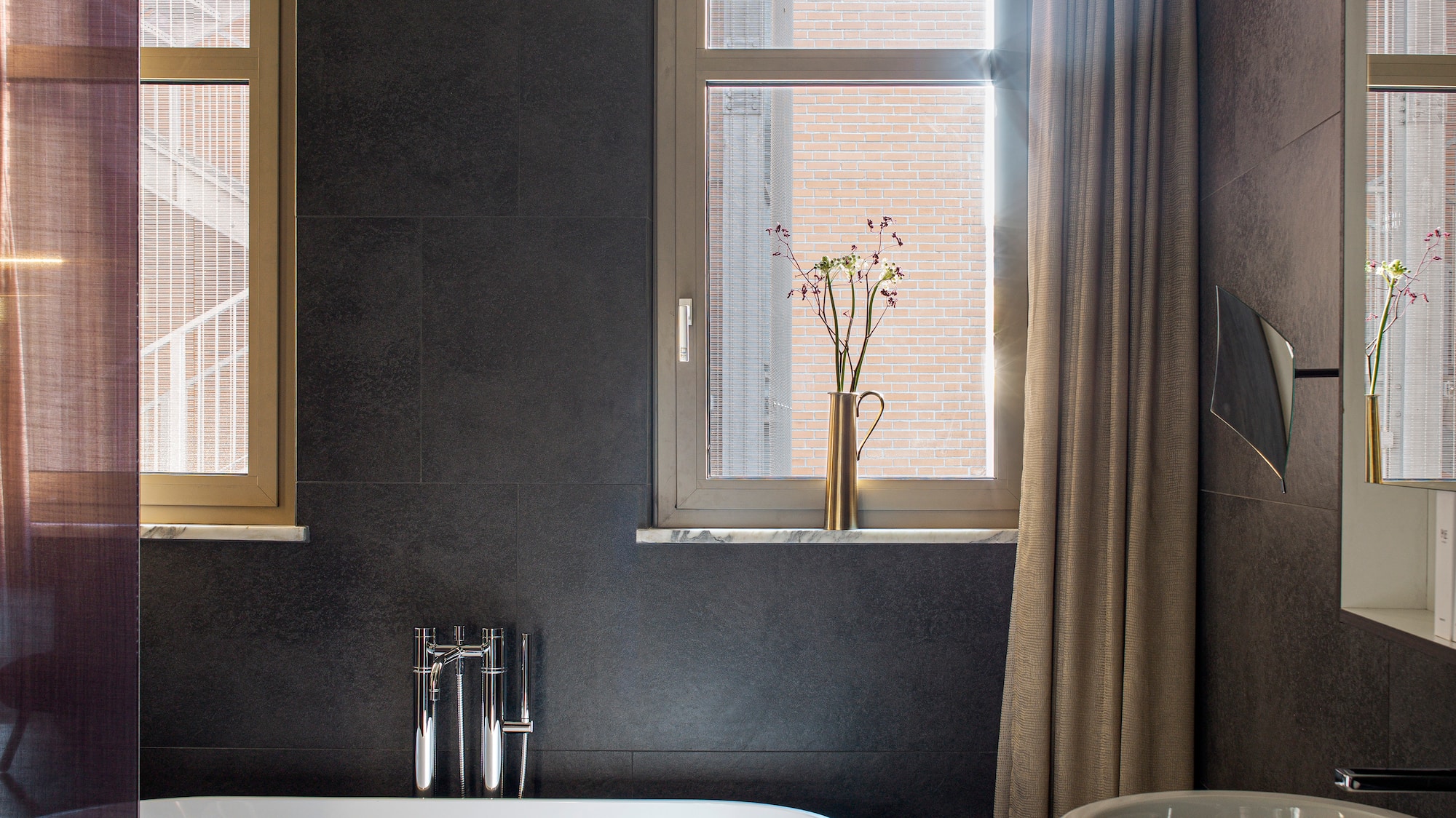 Modern bathroom with freestanding tub, dark wall tiles, basin, and sunlit windows.