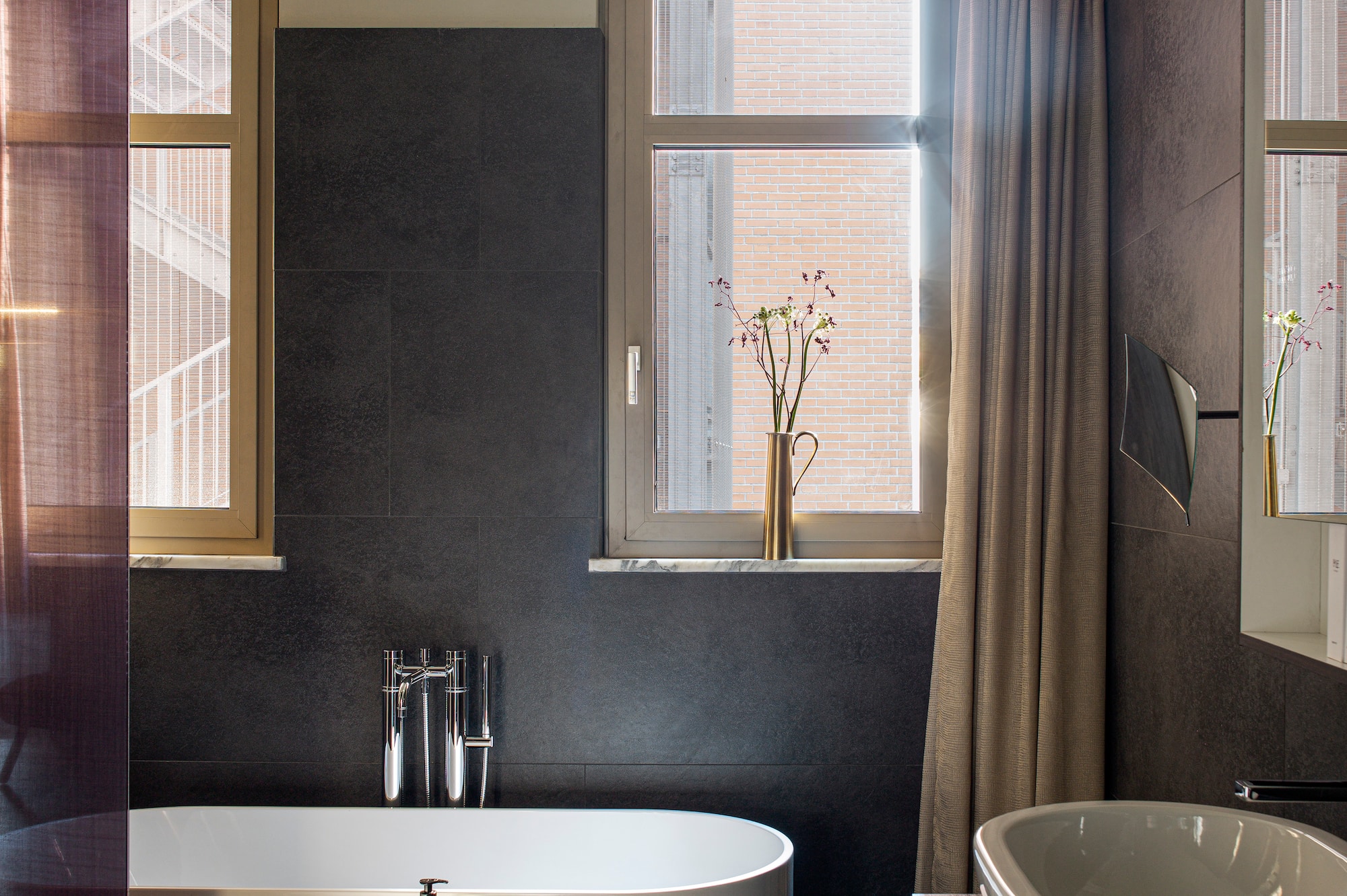 Modern bathroom with freestanding tub, dark wall tiles, basin, and sunlit windows.
