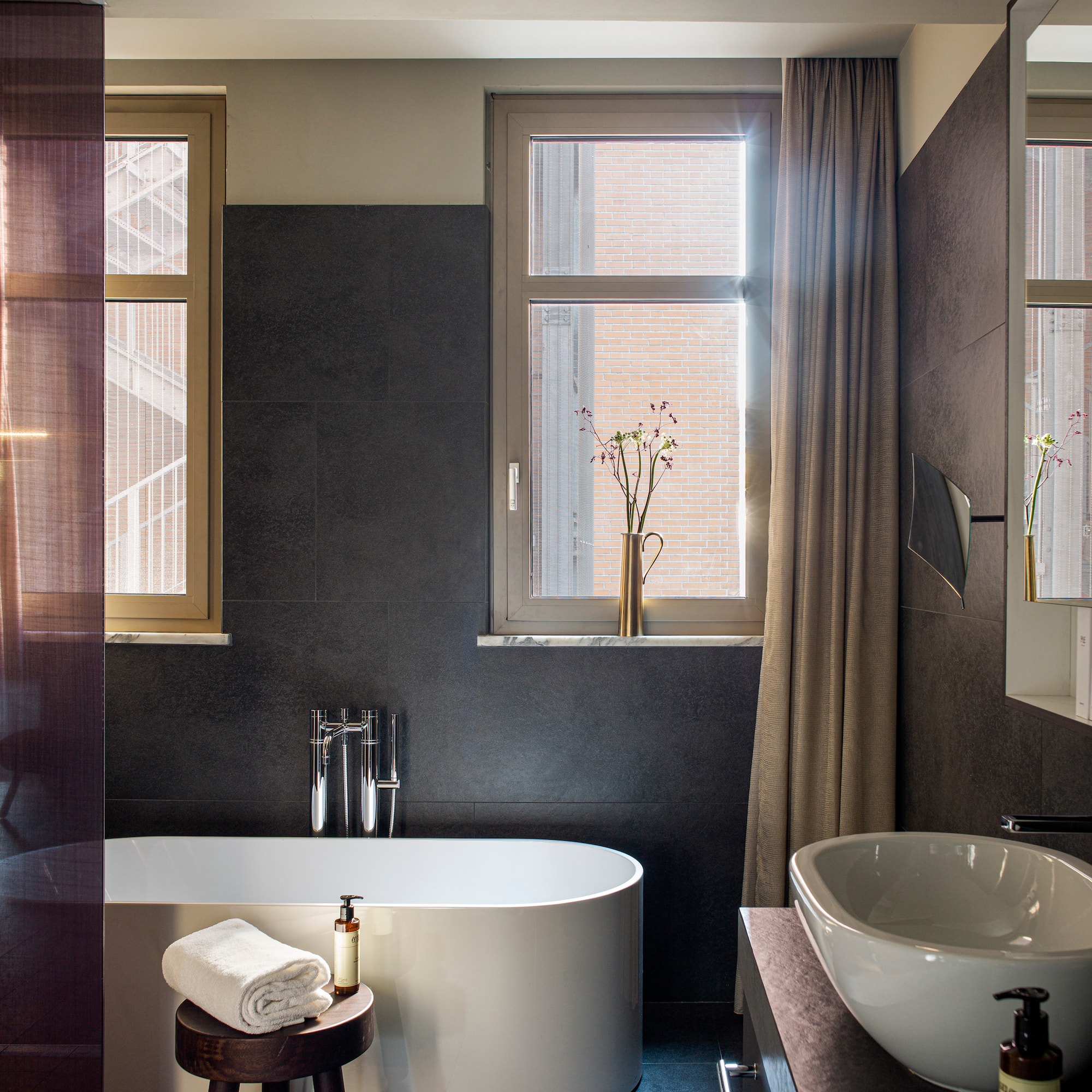 Modern bathroom with freestanding tub, dark wall tiles, basin, and sunlit windows.