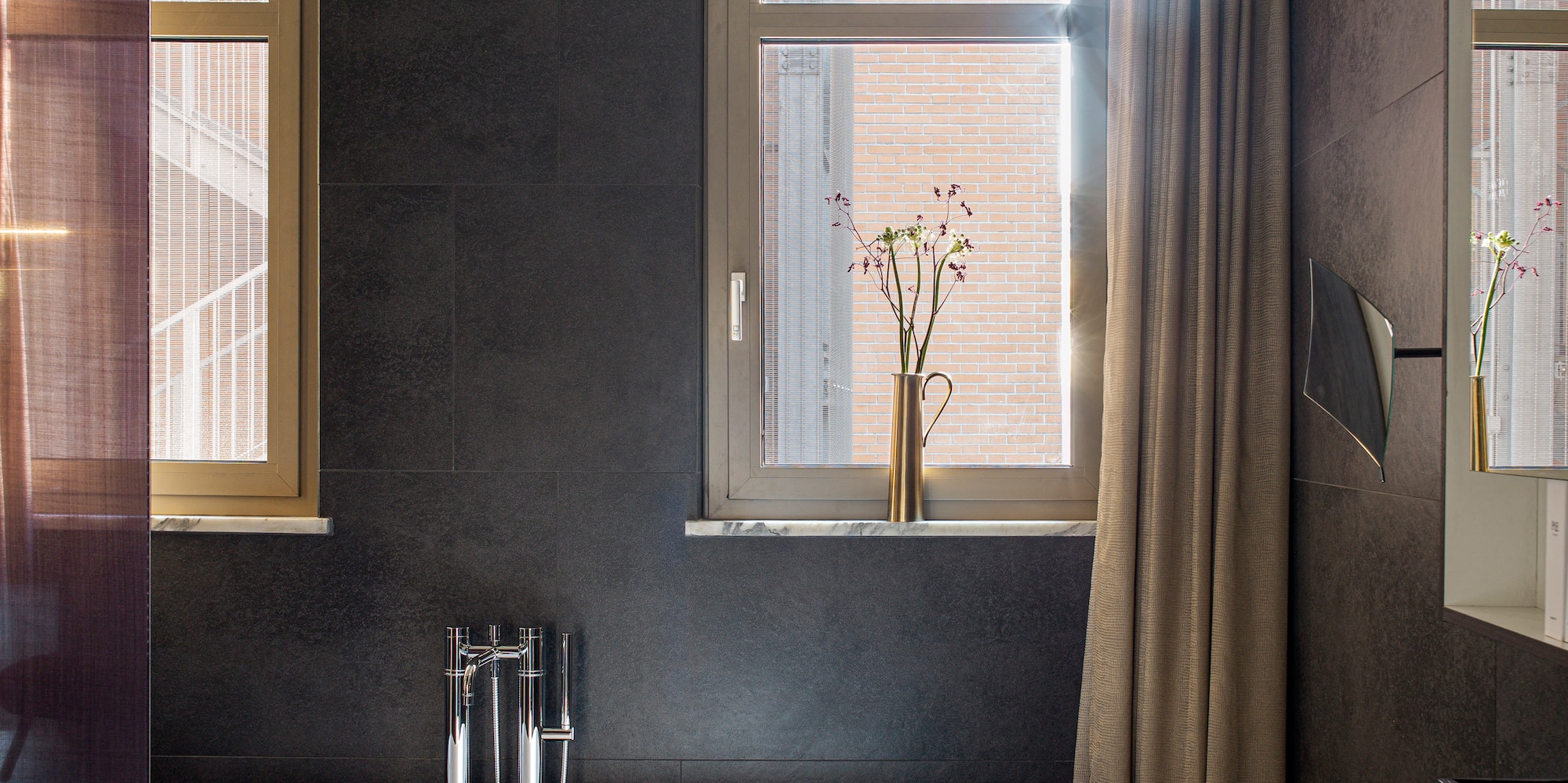 Modern bathroom with freestanding tub, dark wall tiles, basin, and sunlit windows.