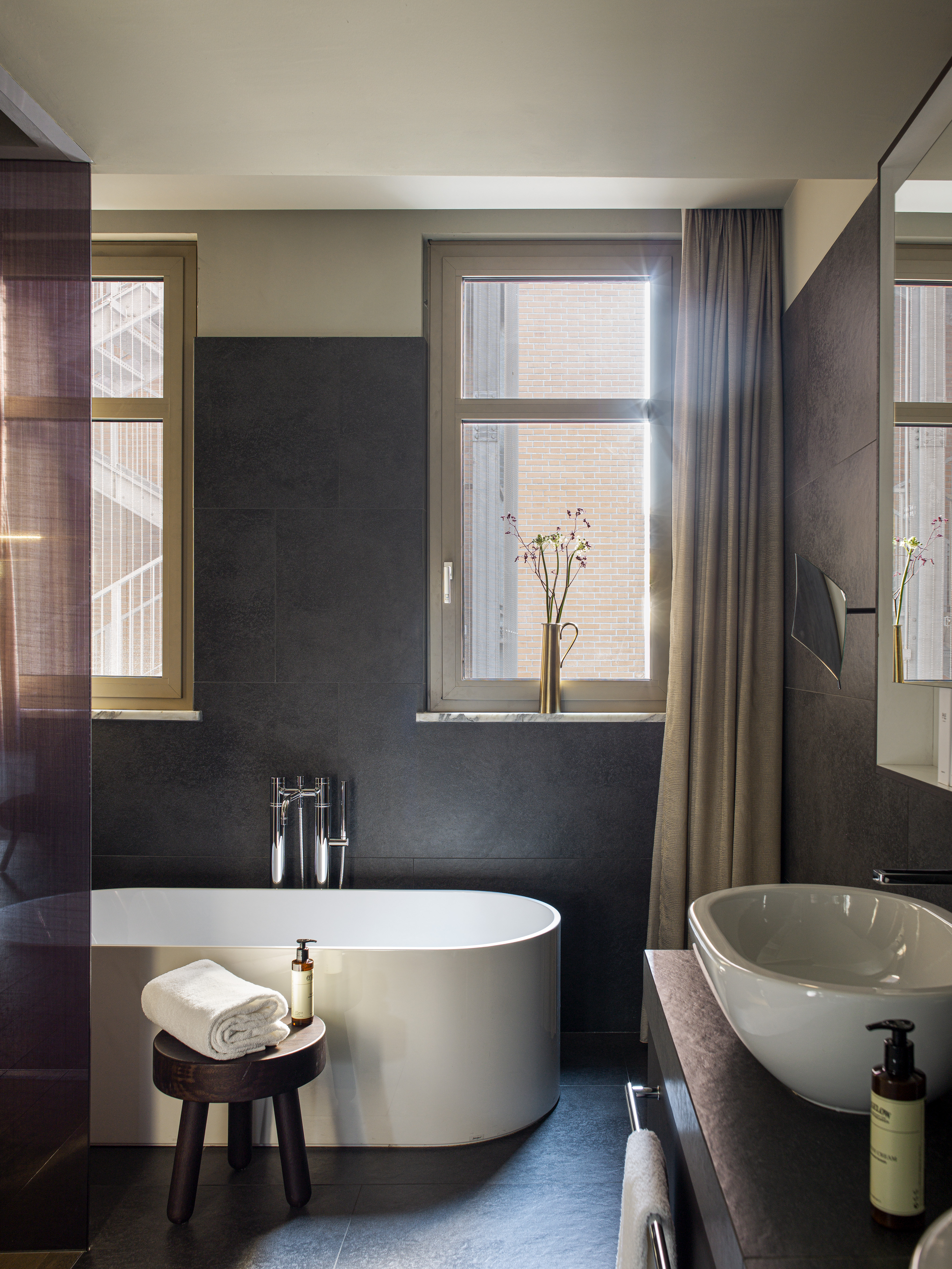 Modern bathroom with freestanding tub, dark wall tiles, basin, and sunlit windows.
