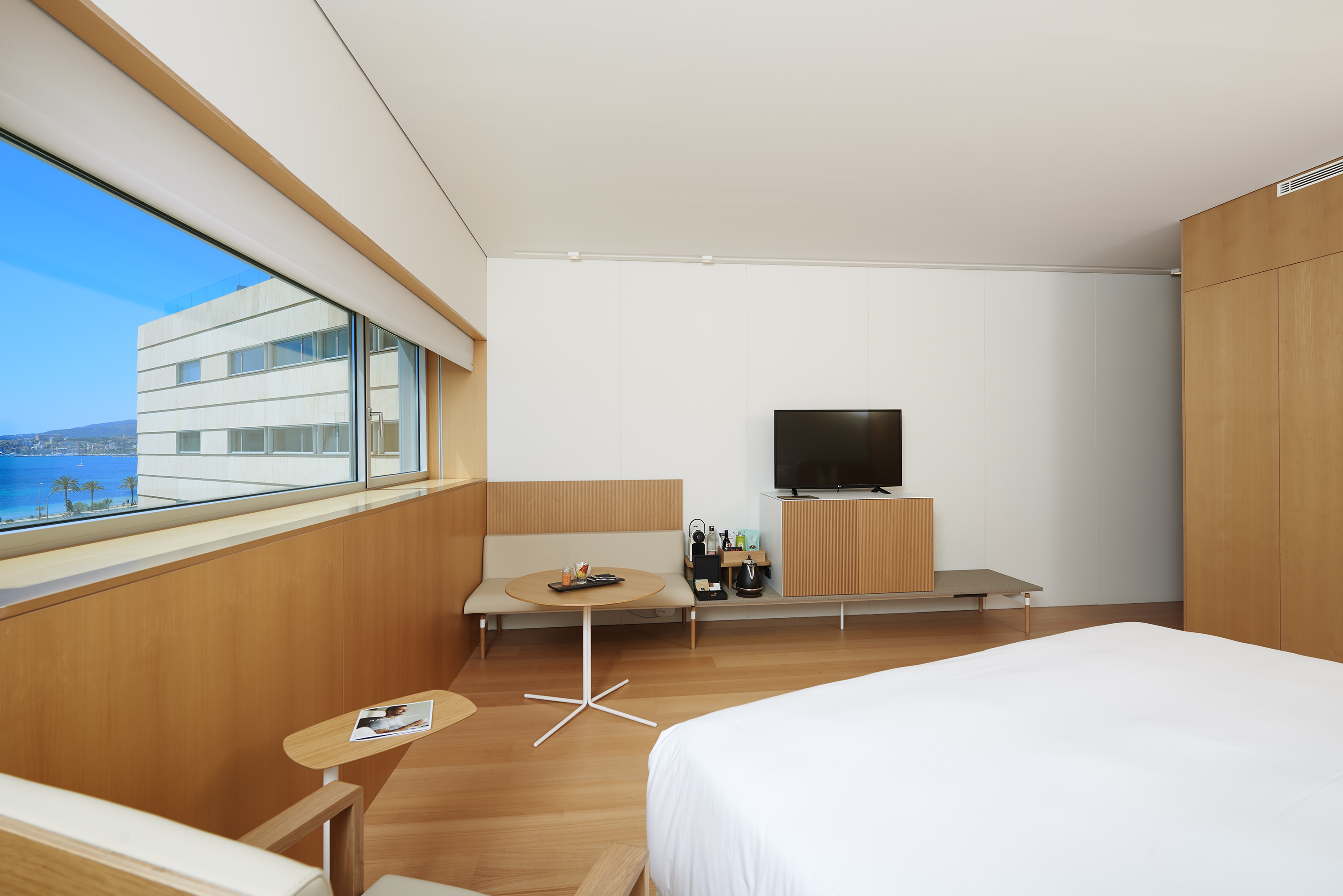 Modern hotel room with ocean view, wooden interior, bed, TV, and large window.
