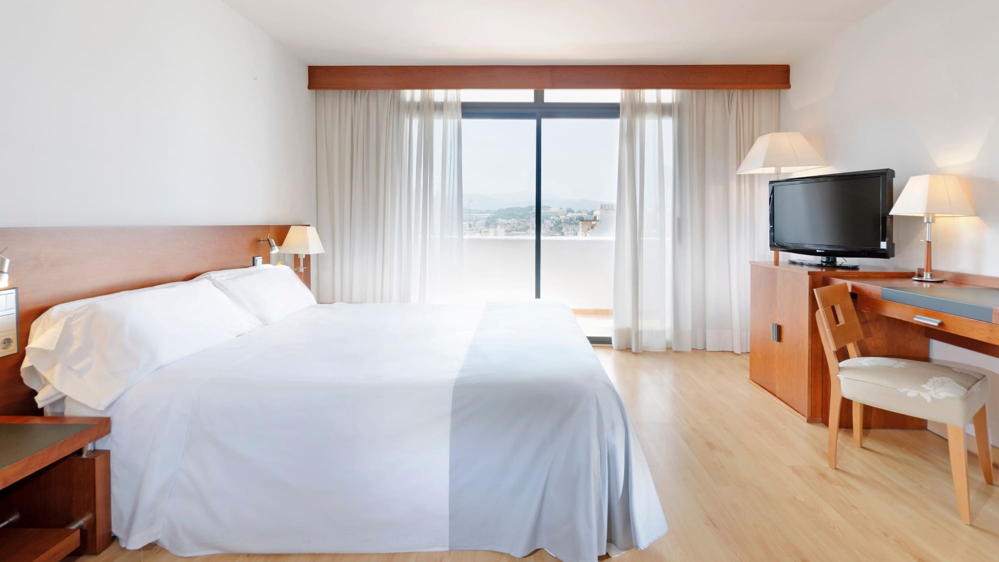 Spacious hotel room with white bed, wooden furniture, TV, and a balcony view.