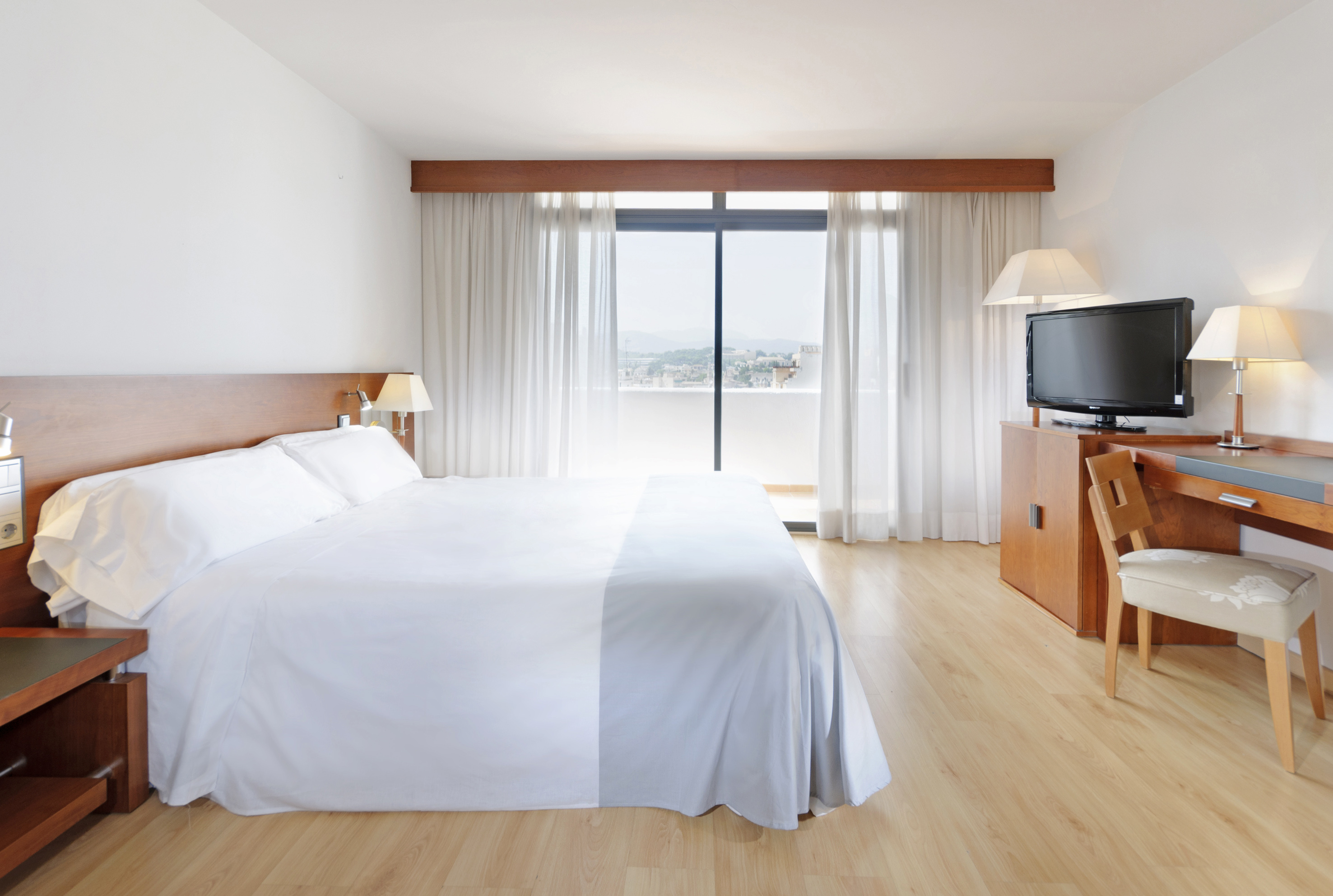 Spacious hotel room with white bed, wooden furniture, TV, and a balcony view.