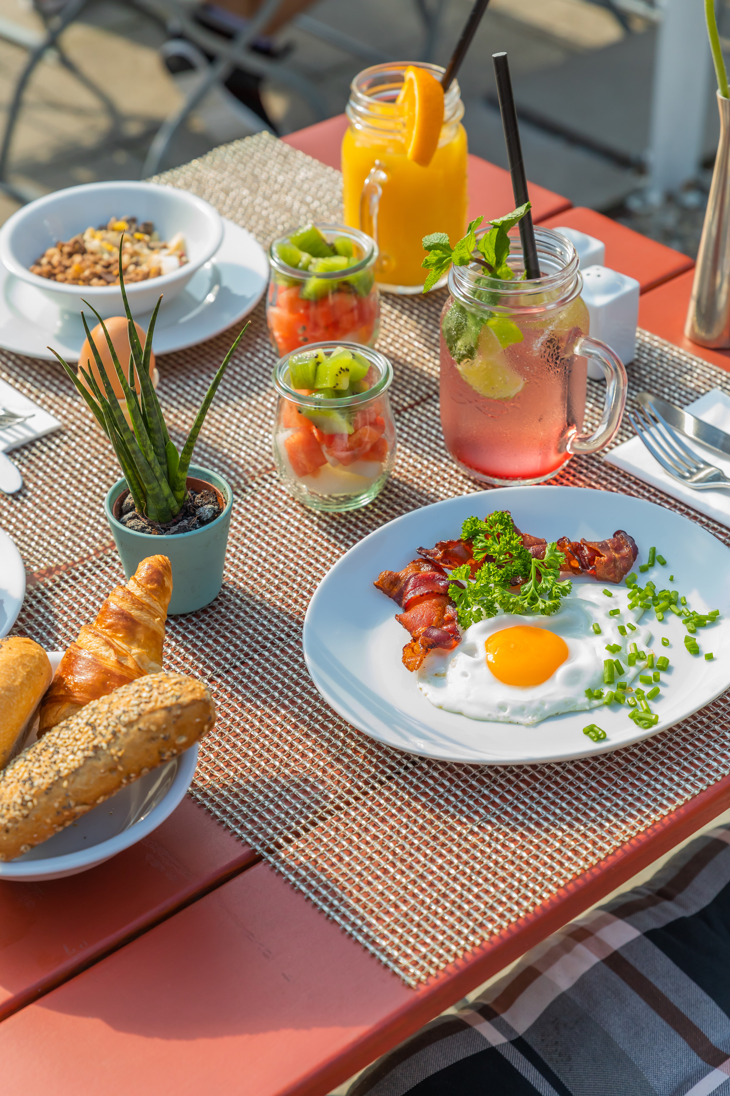 Sunny outdoor breakfast with fried egg, bacon, fruit, muesli, juice, croissants.