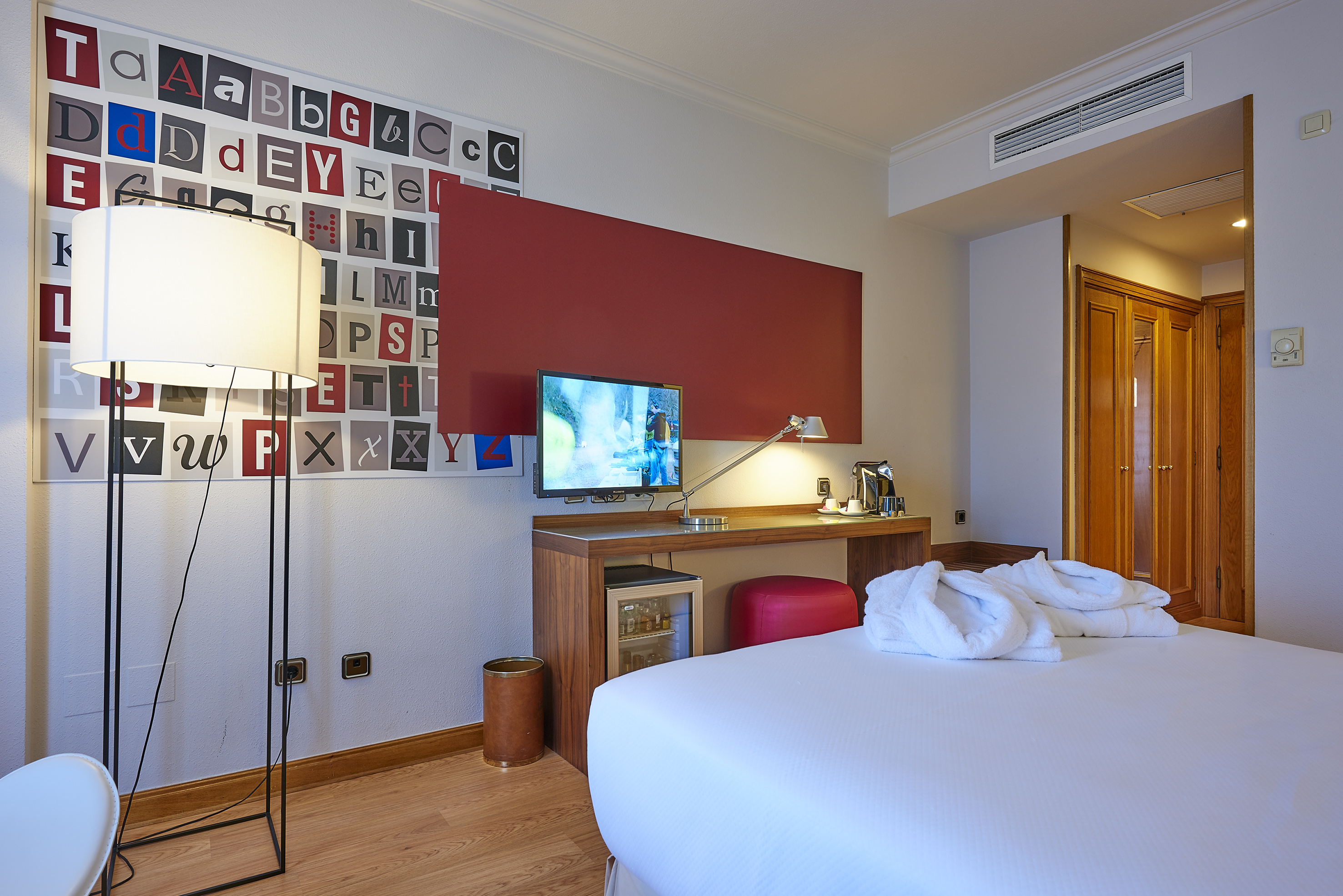 Modern hotel room with white bed, TV, desk, and colorful alphabet wall art.