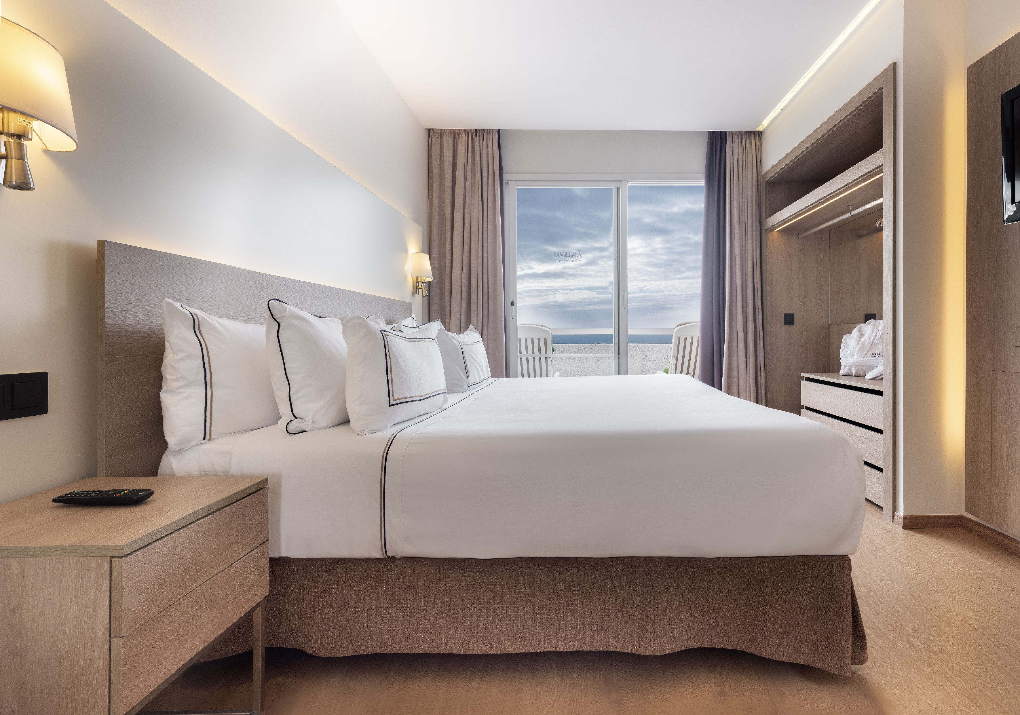 Modern hotel bedroom with large bed, wooden furniture, and ocean view balcony.