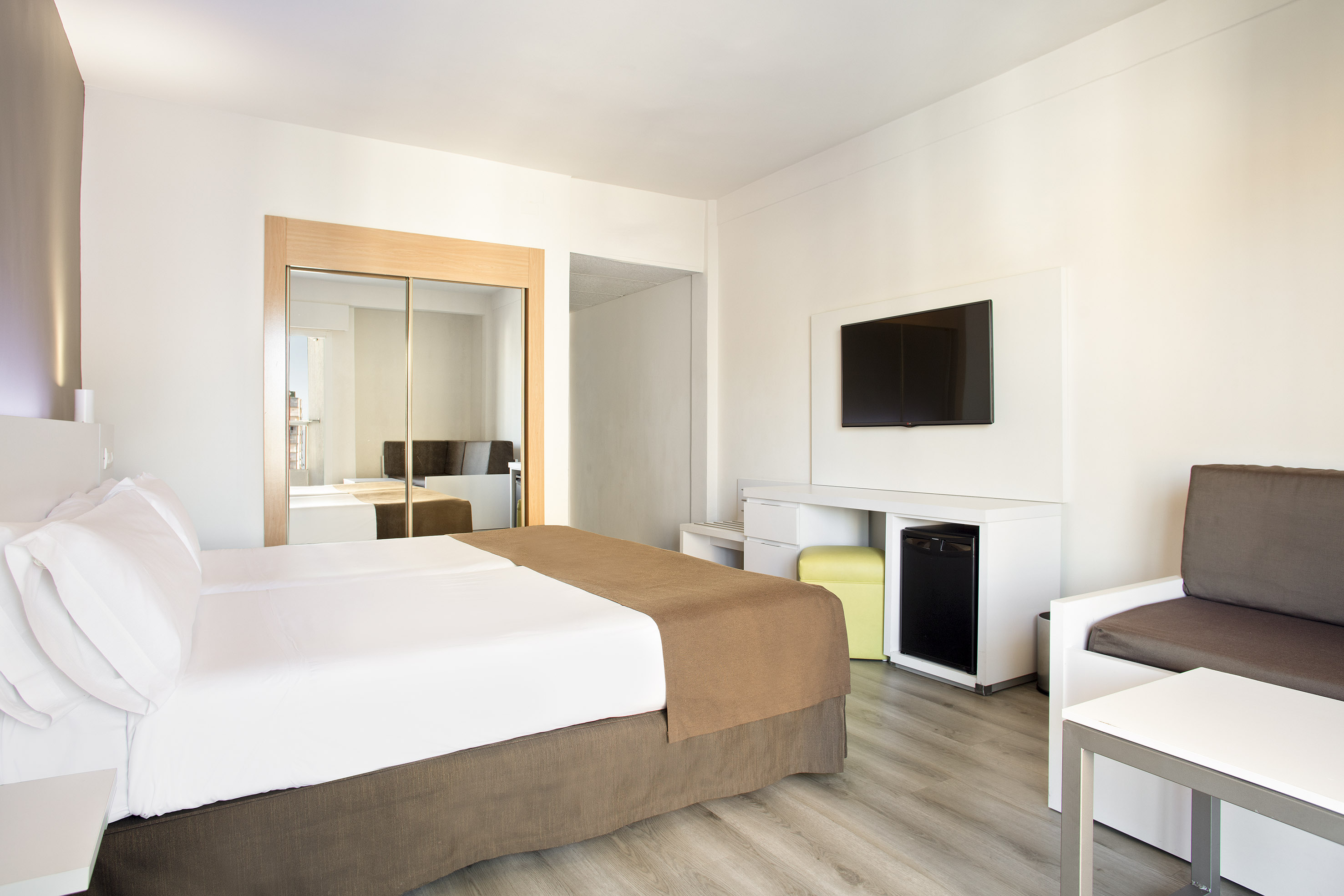 Bright modern hotel room with white bed, mirrored wardrobe, TV, and grey sofa.