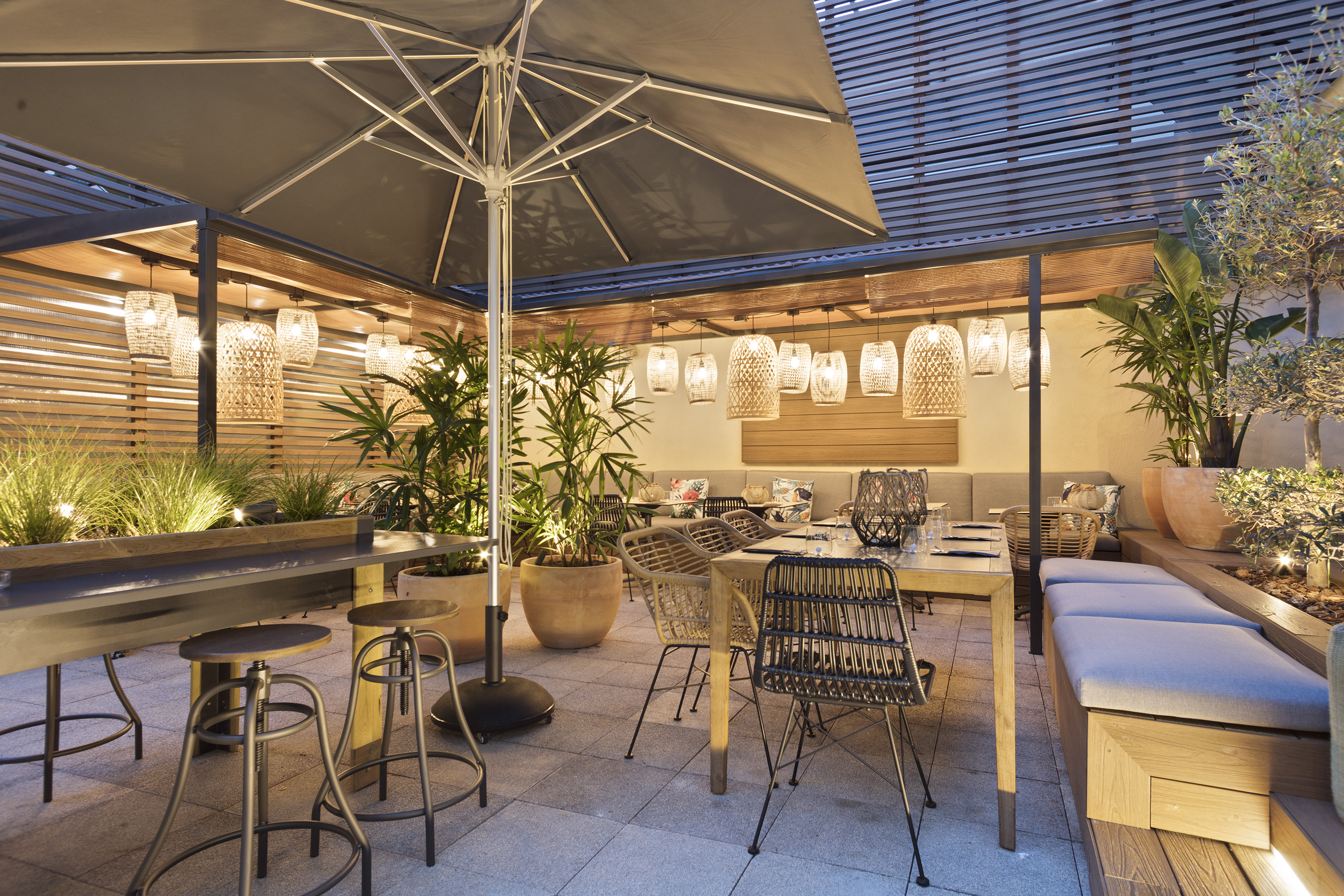 Modern outdoor dining terrace with illuminated rattan lamps and green plants.