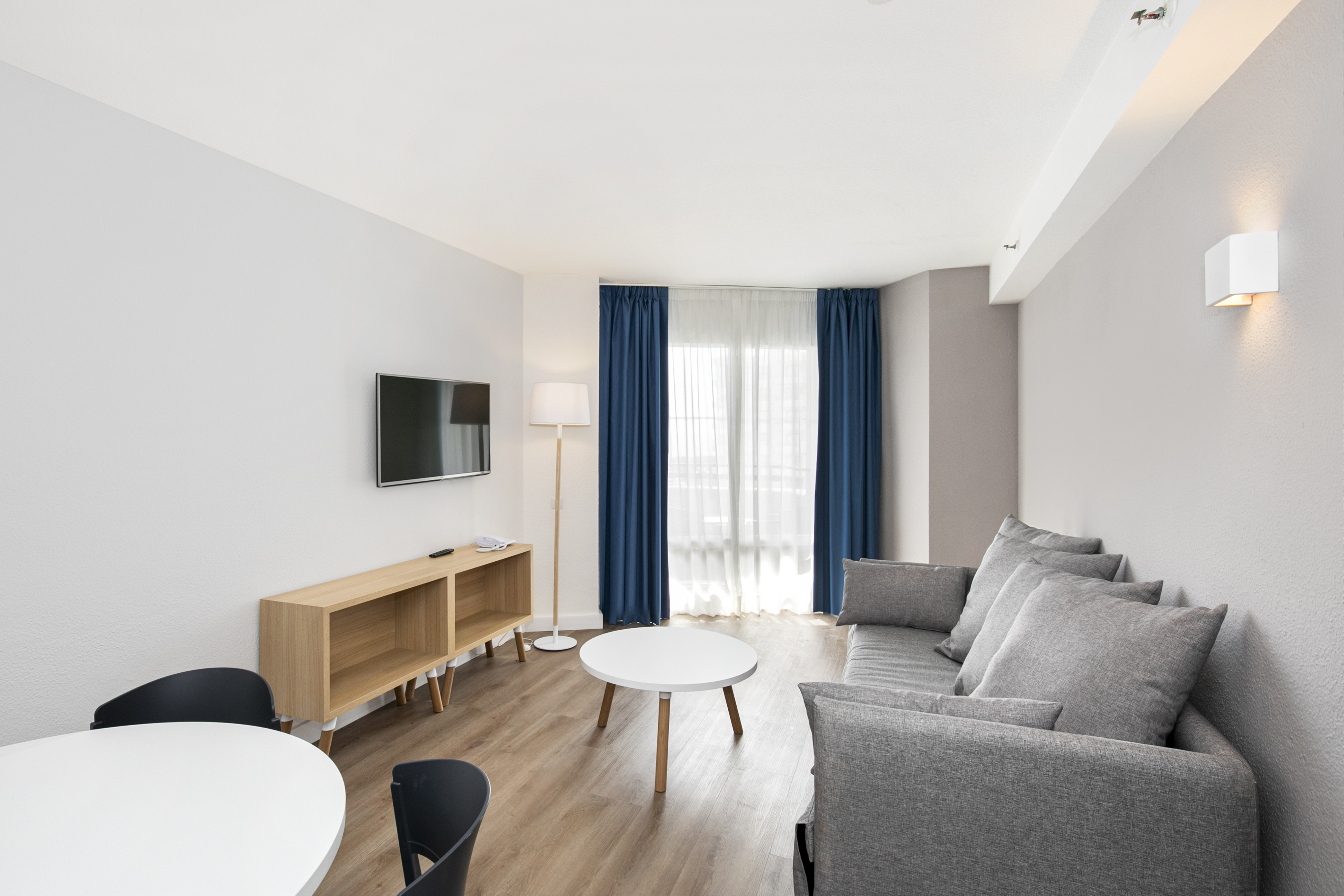 Modern living room with gray sofa, flat-screen TV, wood floor, blue curtains.