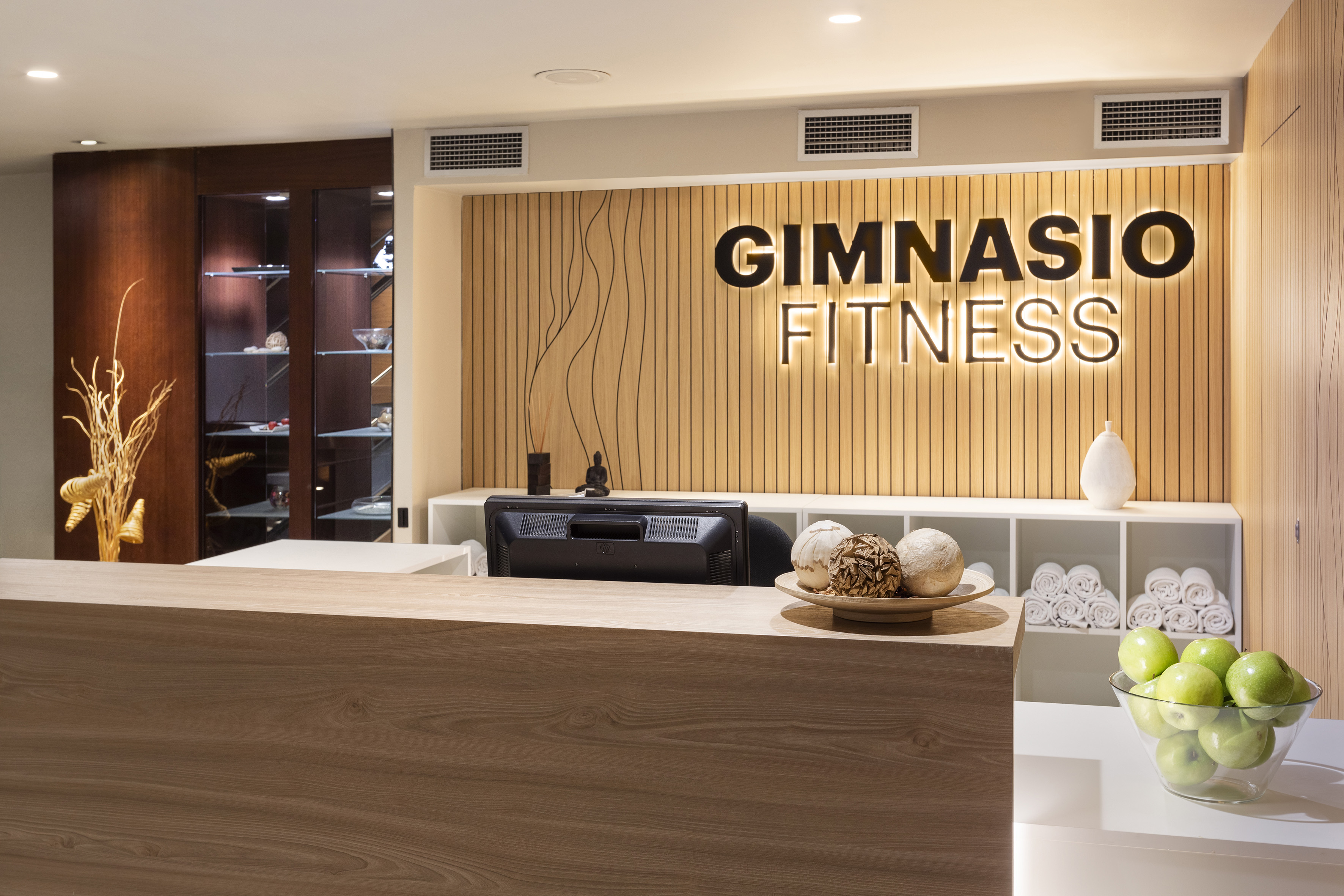 Modern gym reception desk with "Gimnasio Fitness" sign, rolled towels, and green apples.