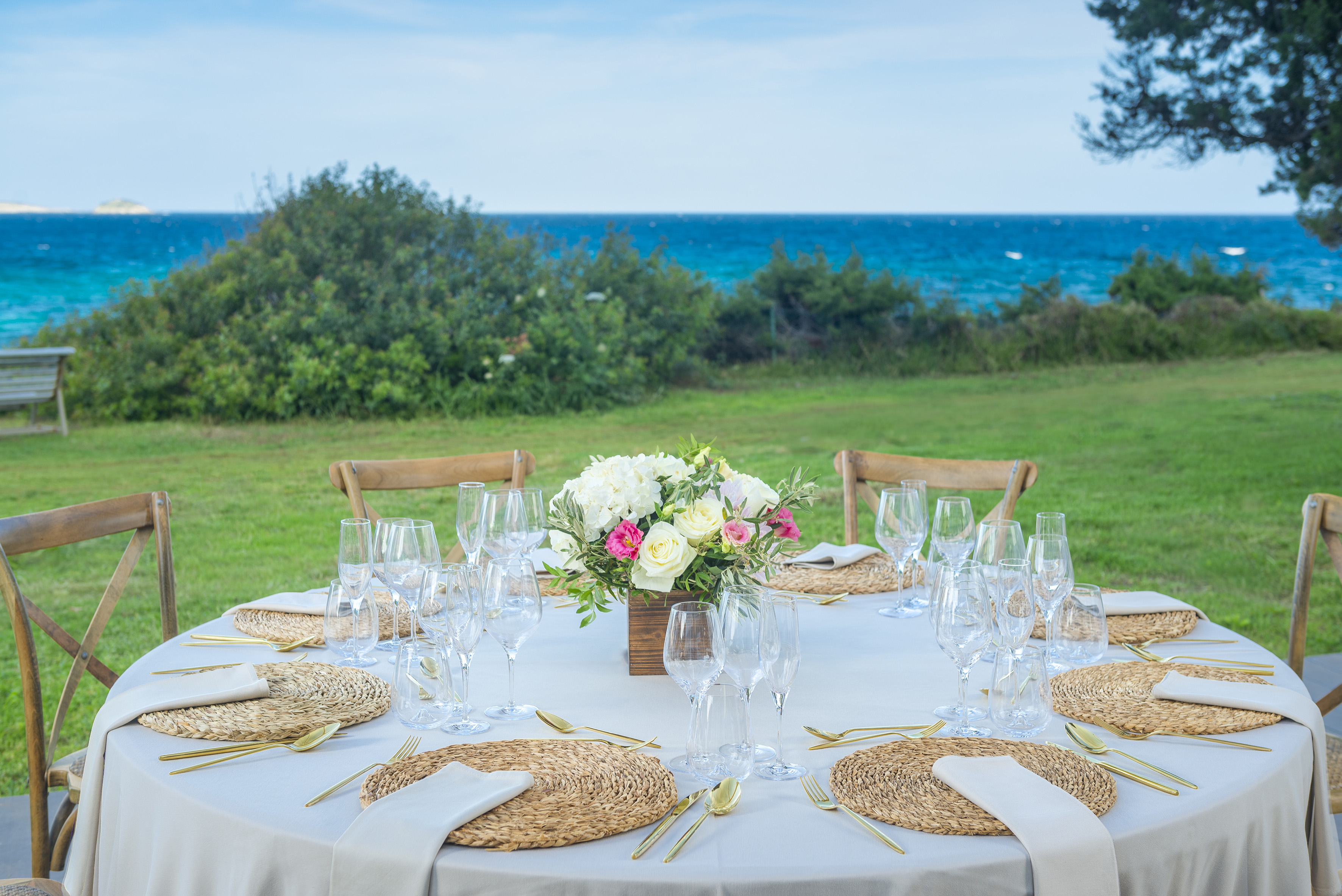 Seaside outdoor event table with floral centerpiece and golden cutlery.
