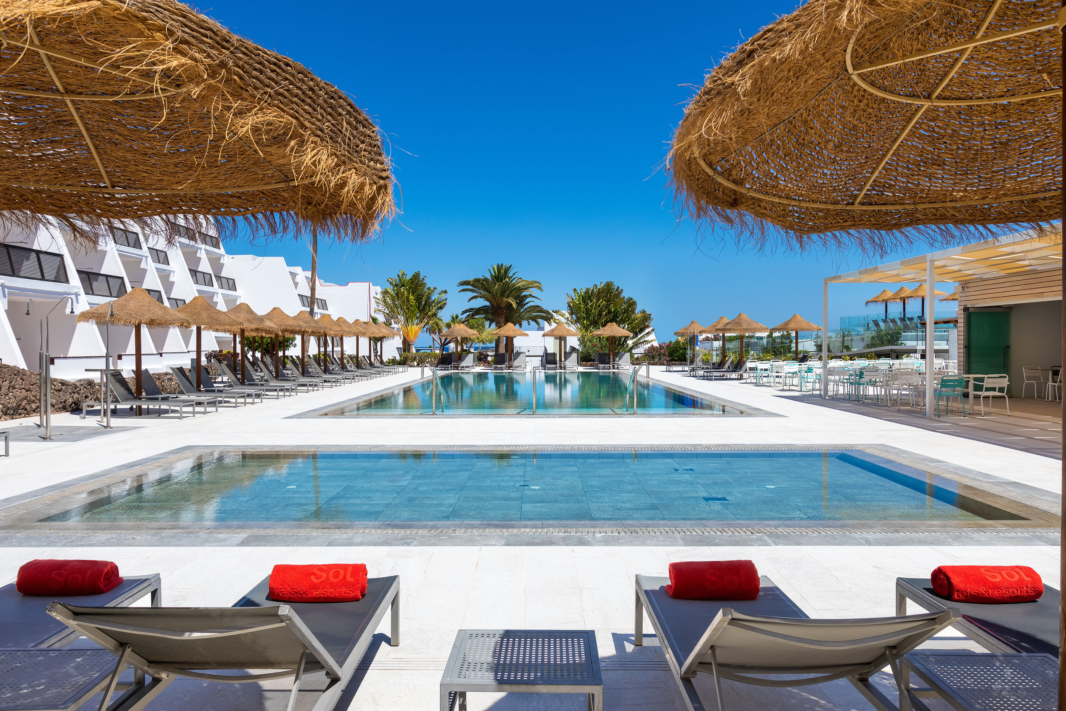 Sunny hotel resort pools with sun loungers, straw umbrellas, and palm trees.
