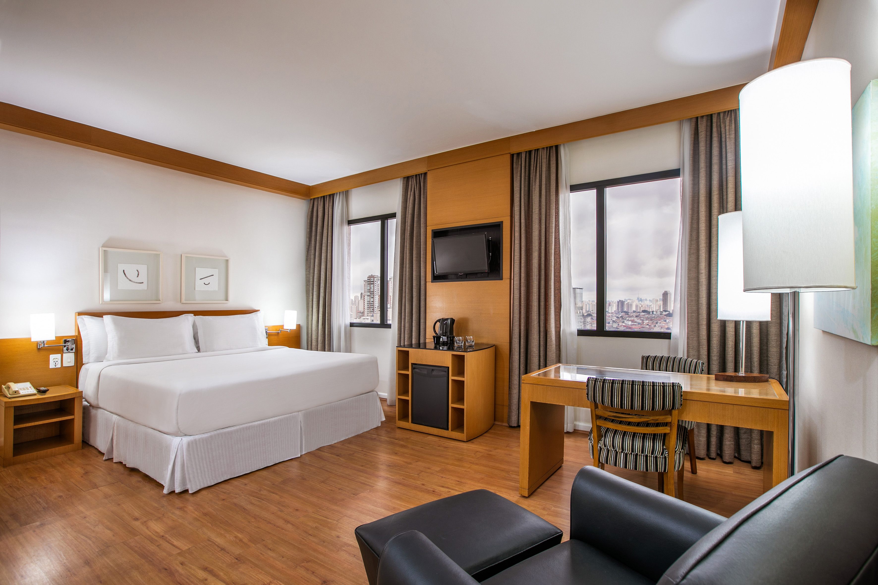 Modern hotel room with king bed, desk, TV, wooden floor, and city view.