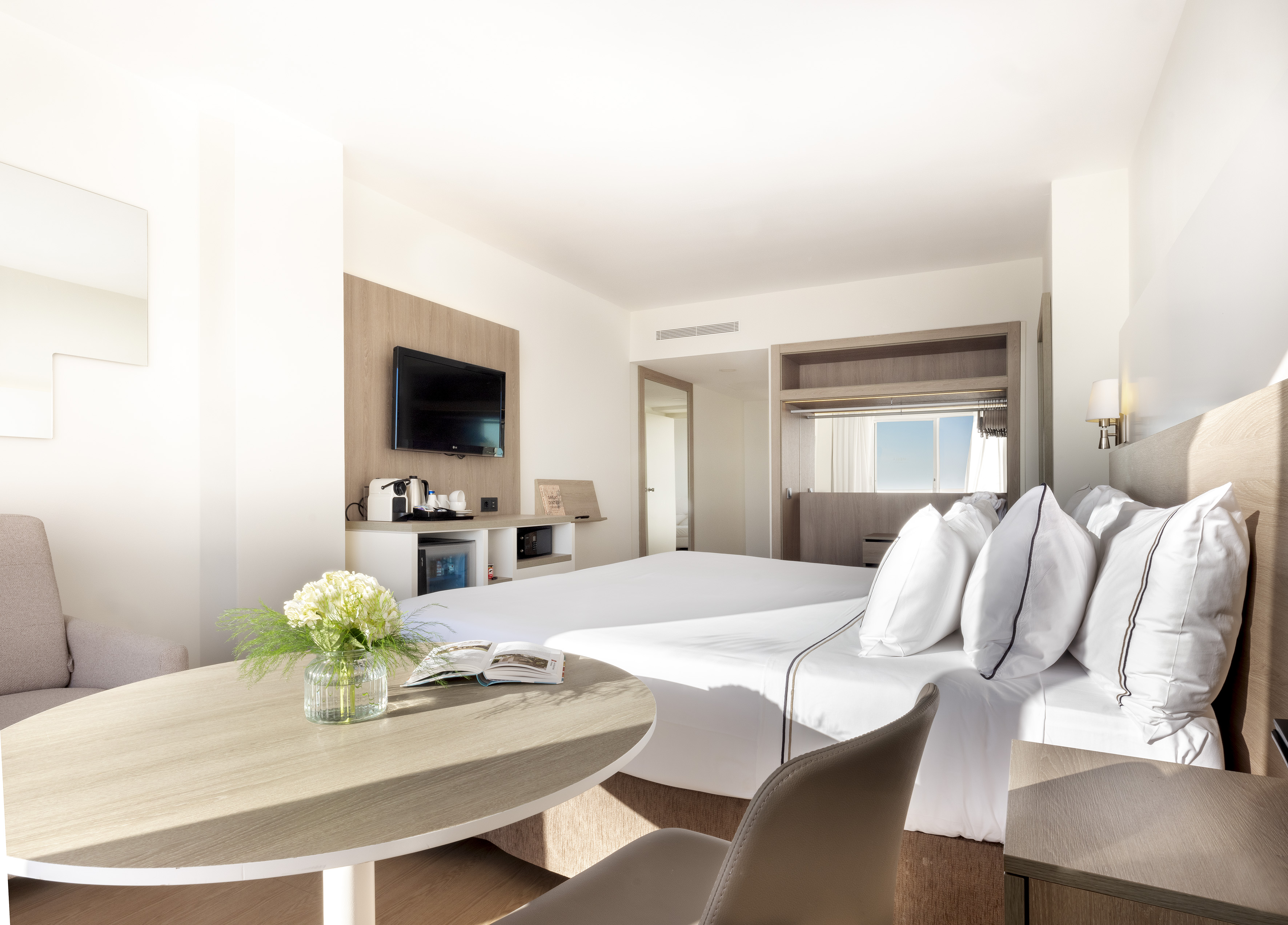 Modern hotel room with two beds, TV, and light wood interior.