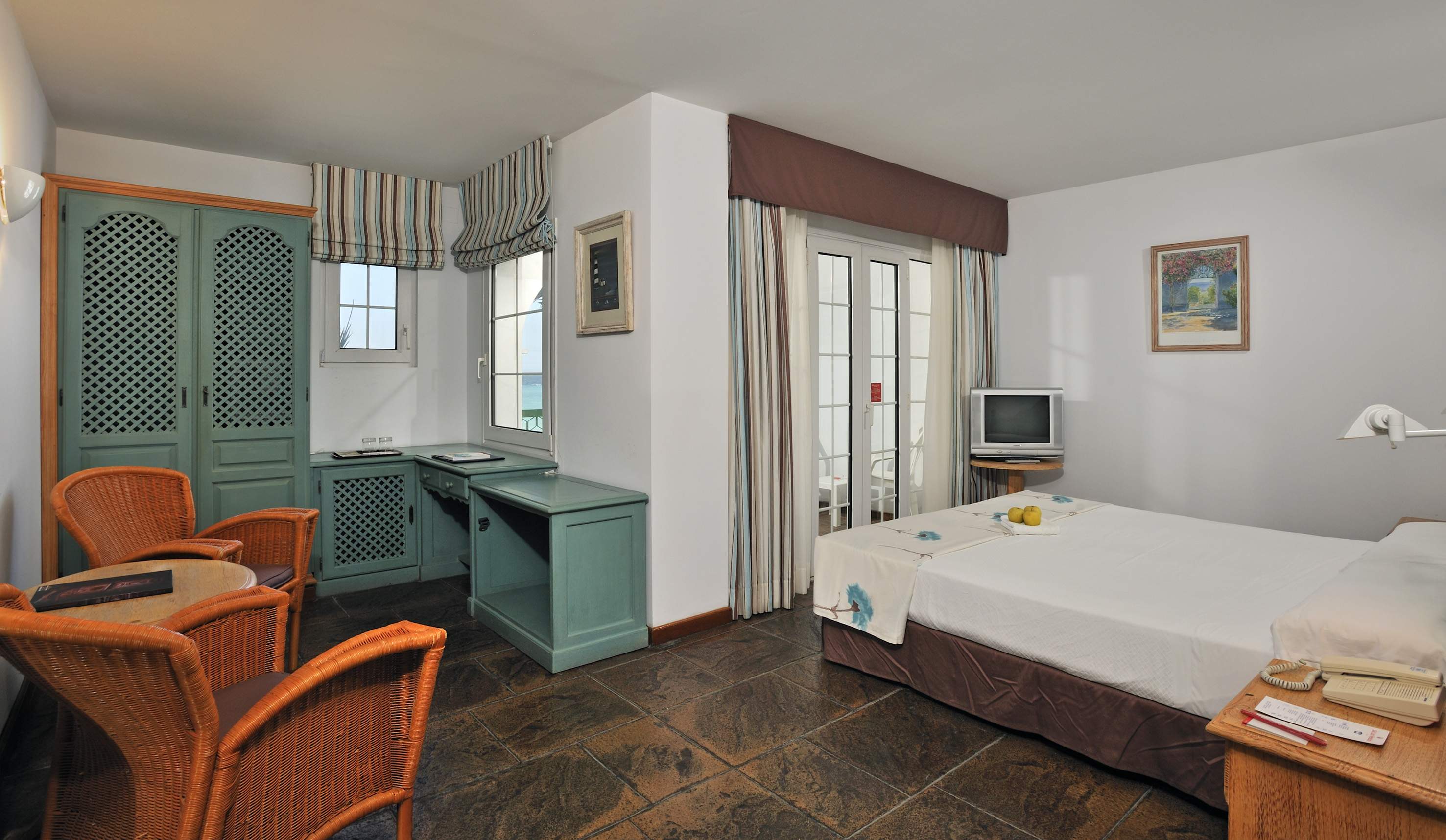 Hotel guest room with bed, teal furniture, wicker chairs, and sea view.