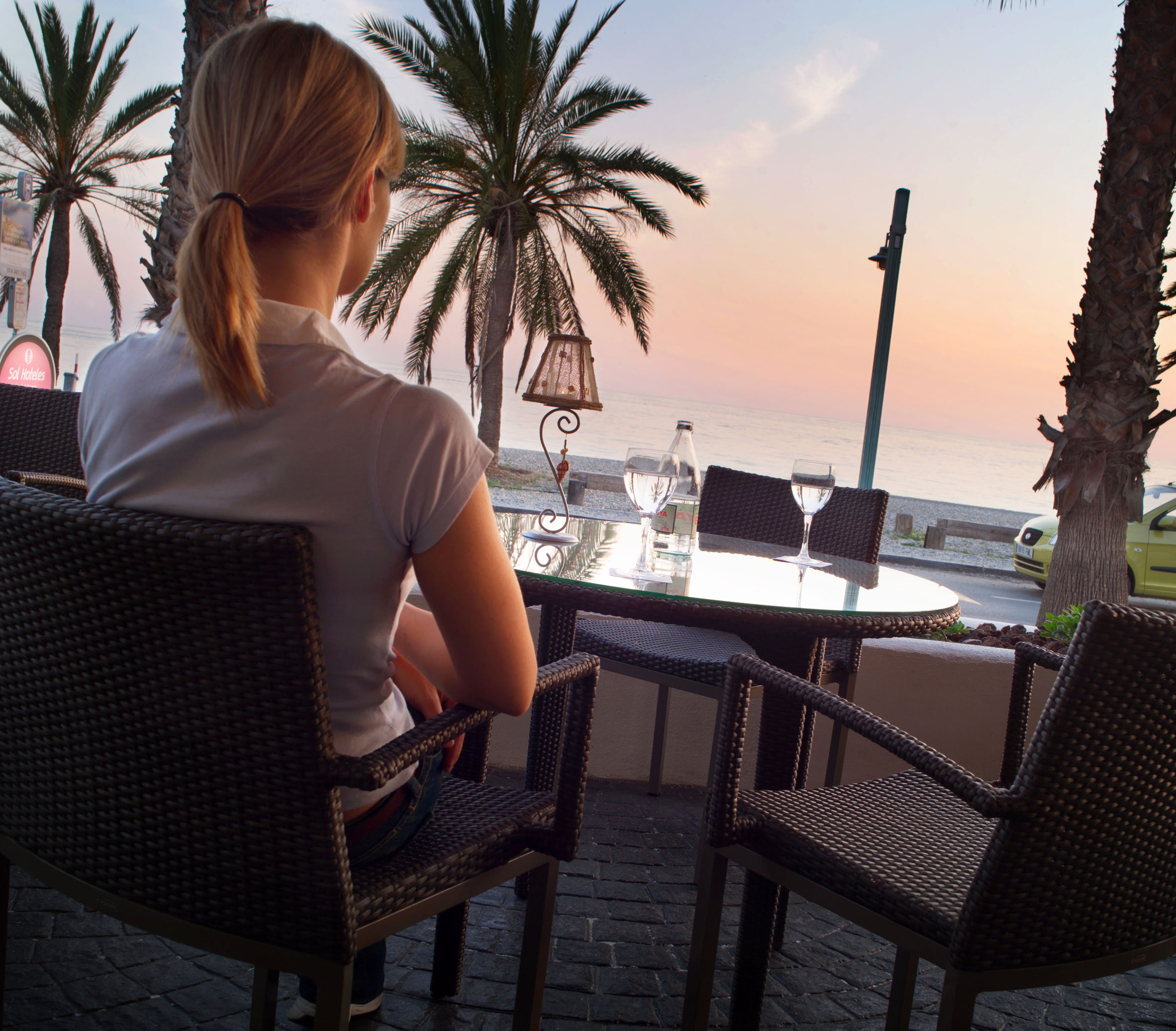 Woman at outdoor beachfront cafe with palm trees and ocean sunset.
