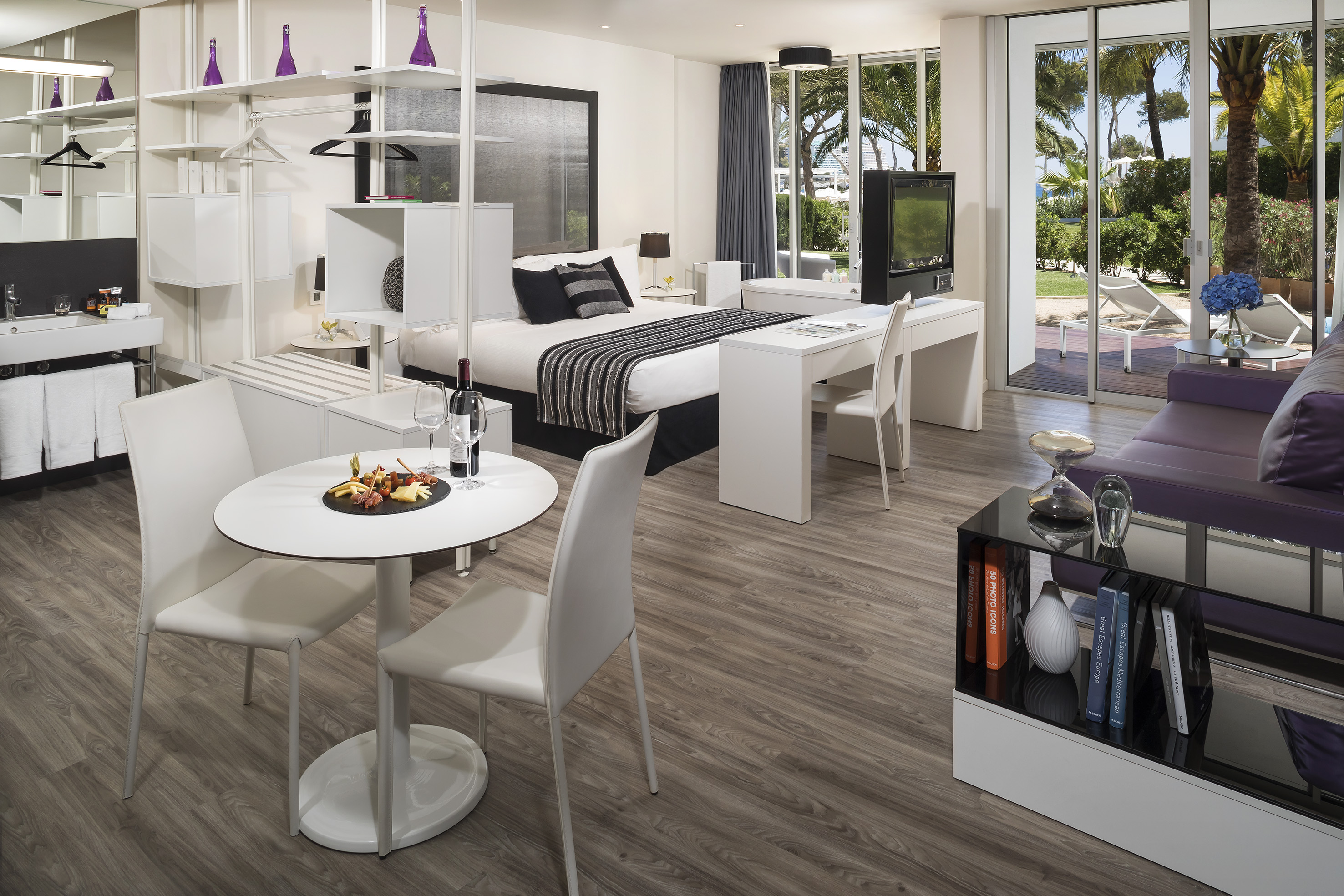 Modern hotel room with bed, purple sofa, dining area, and tropical garden view.