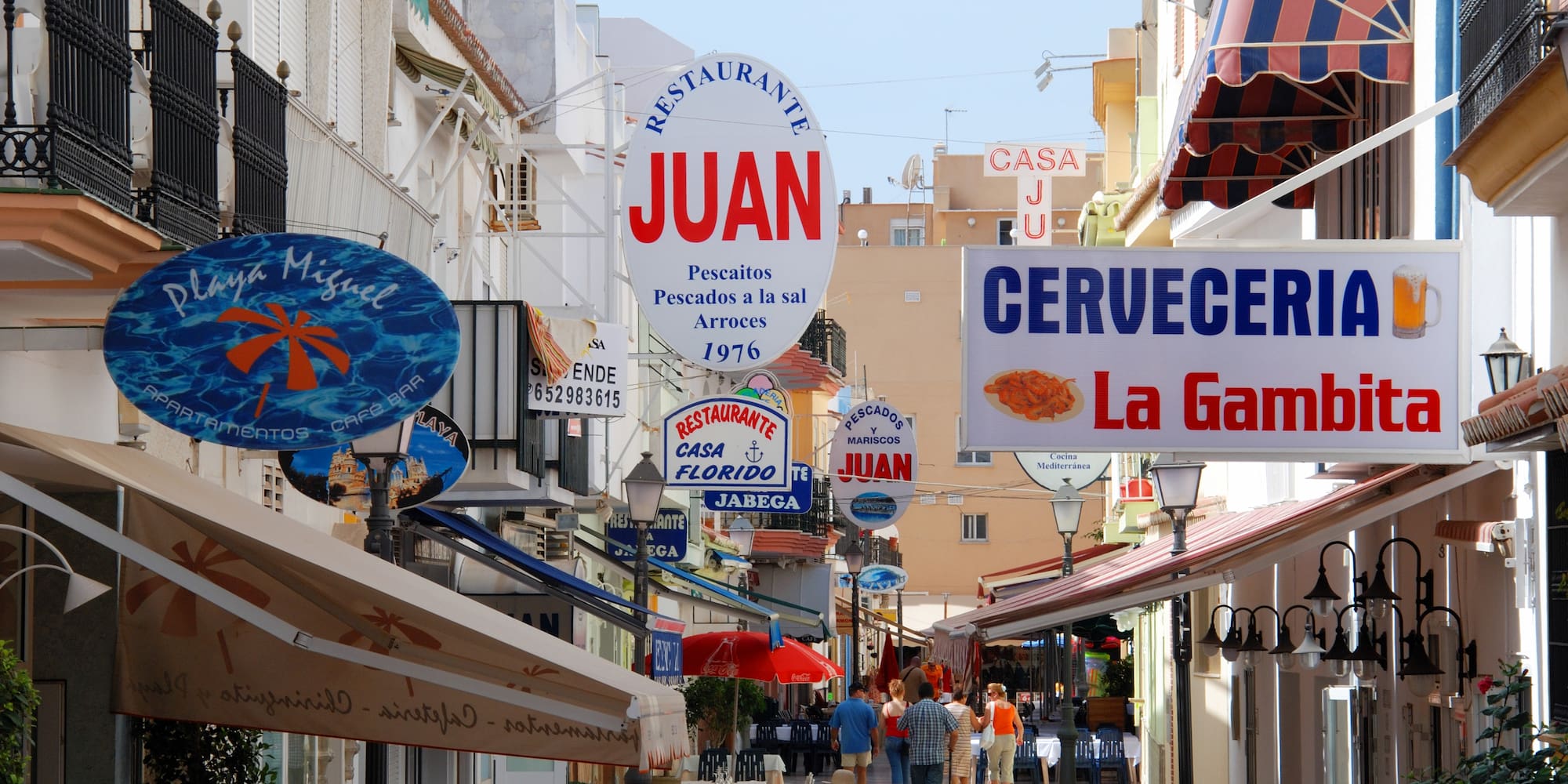 Spanish street with restaurant signs, outdoor dining, and pedestrians.