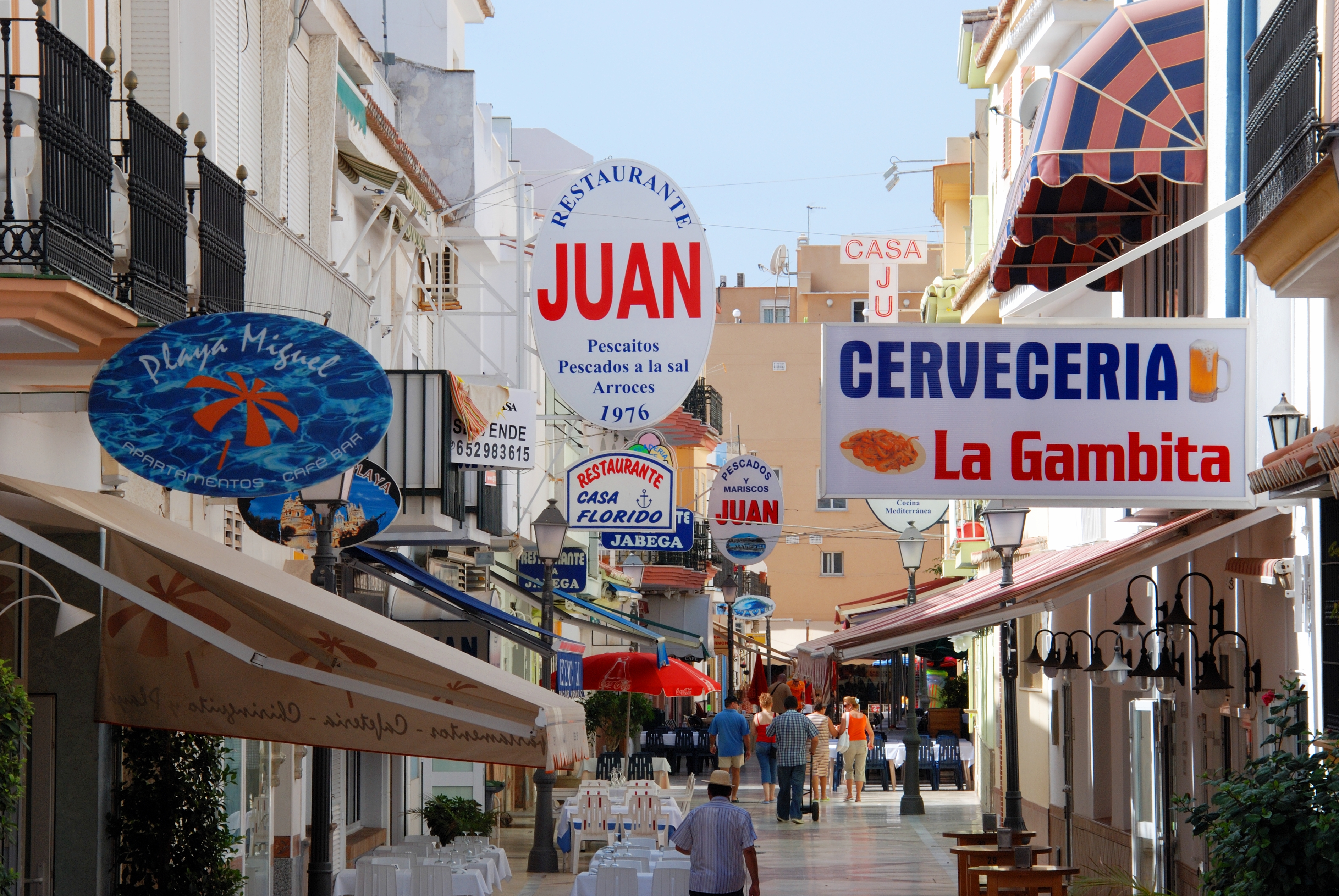 Vibrant Mediterranean street with restaurant signs, outdoor dining, and people.