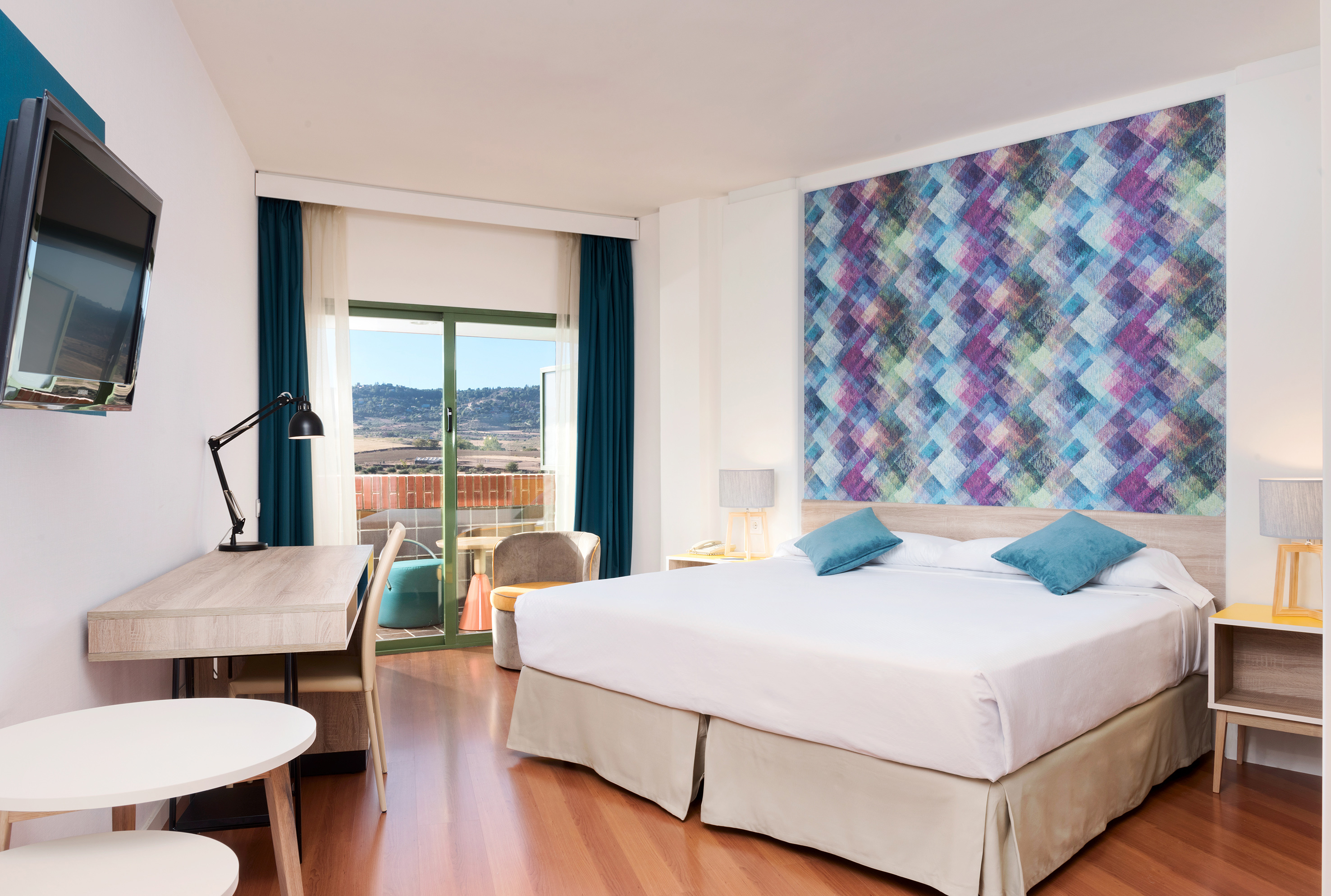 Modern hotel room with bed, geometric accent wall, desk, and balcony mountain view.
