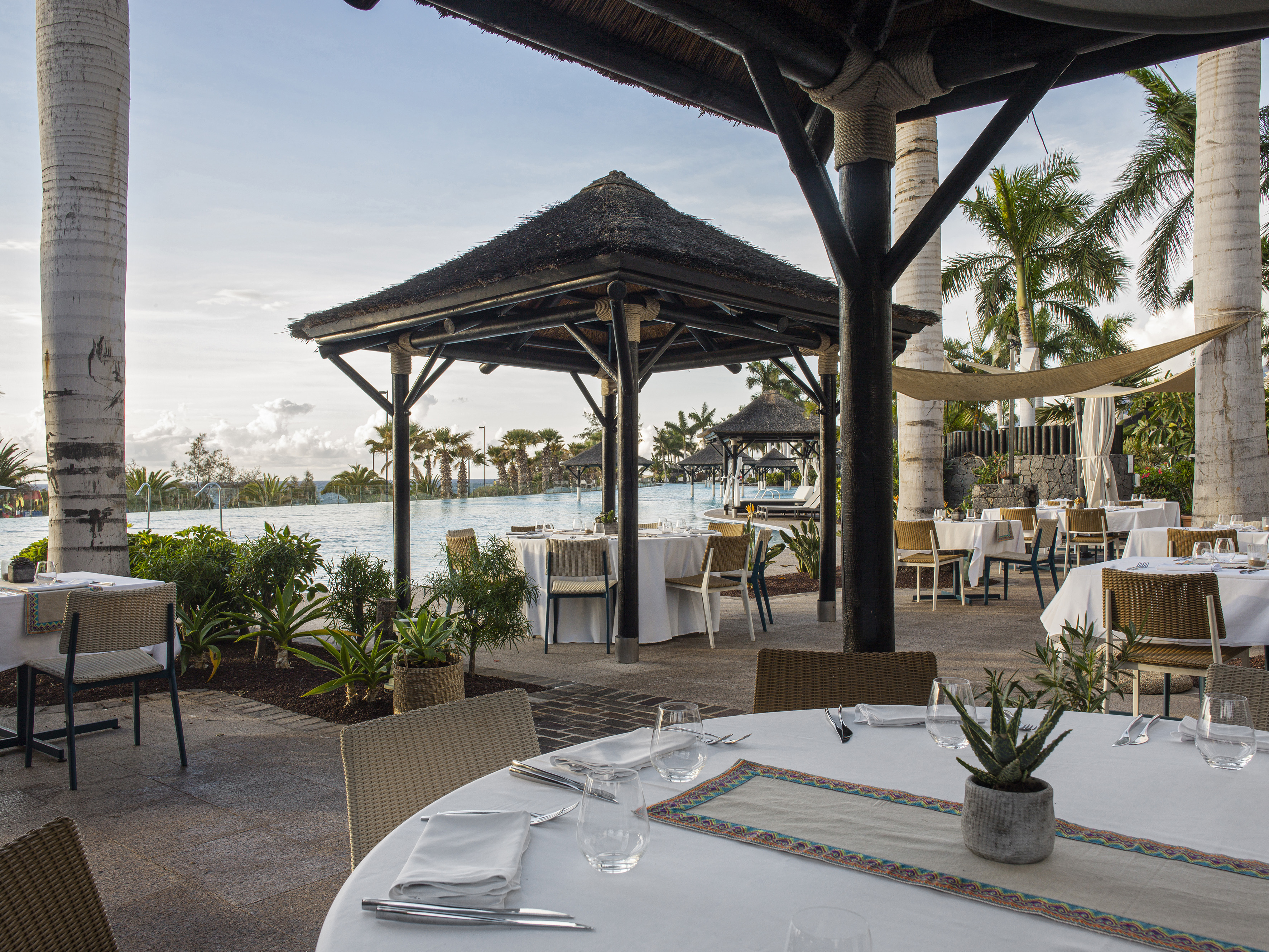 Elegant outdoor restaurant with thatched roofs, palm trees, and infinity pool.