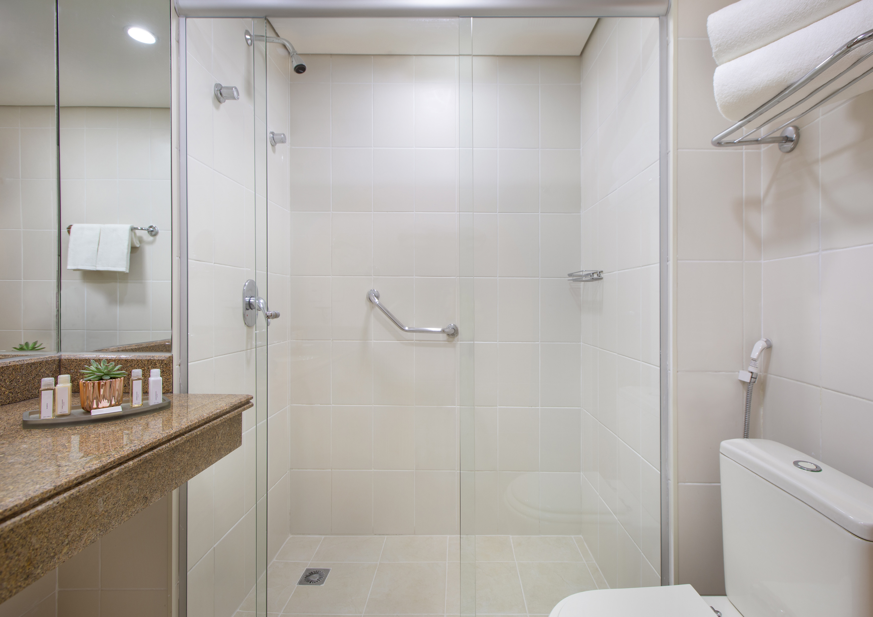Modern hotel bathroom with glass shower, white tiles, granite counter, and toilet.