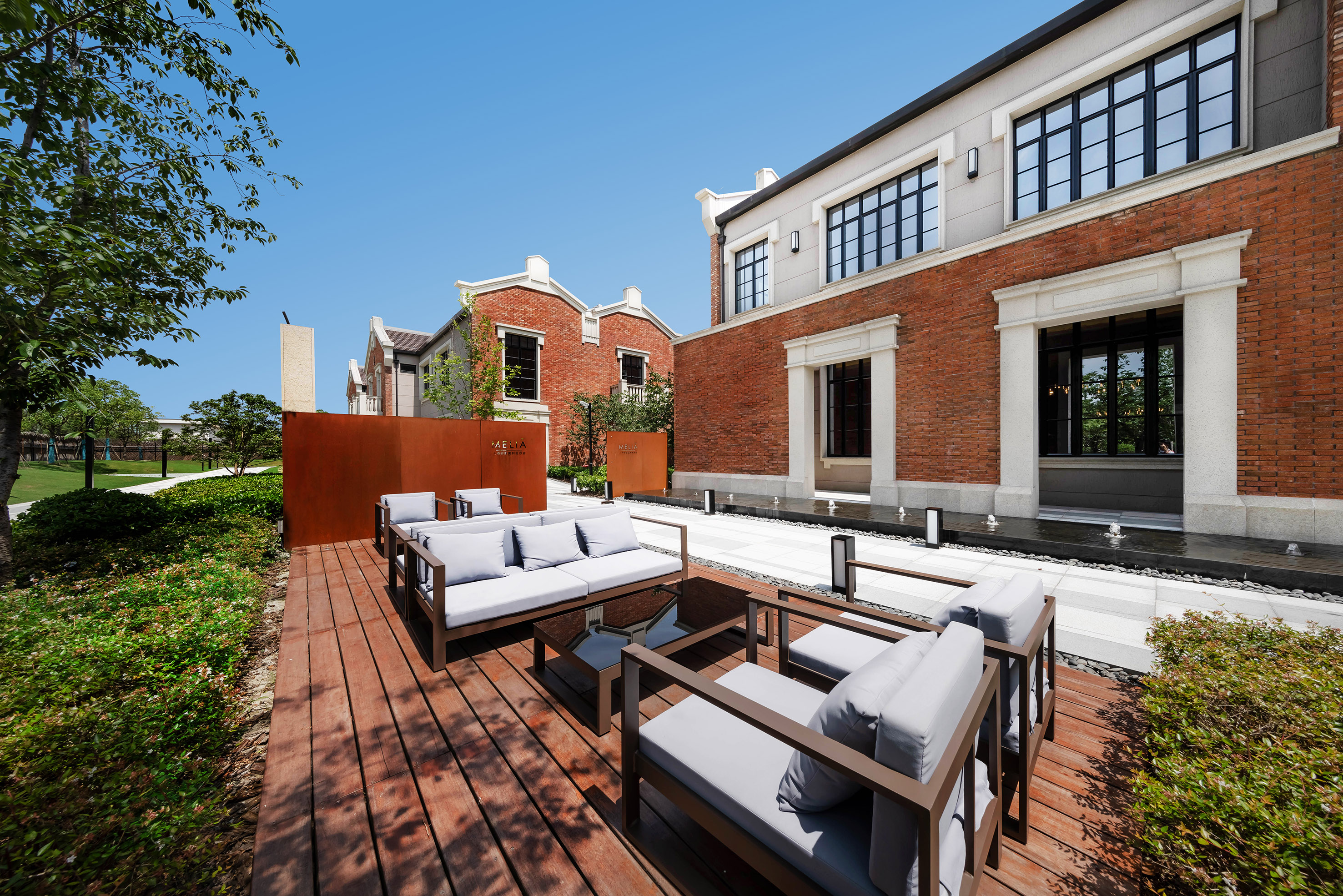 Modern outdoor lounge on a wooden deck, brick villas, and green landscaping.