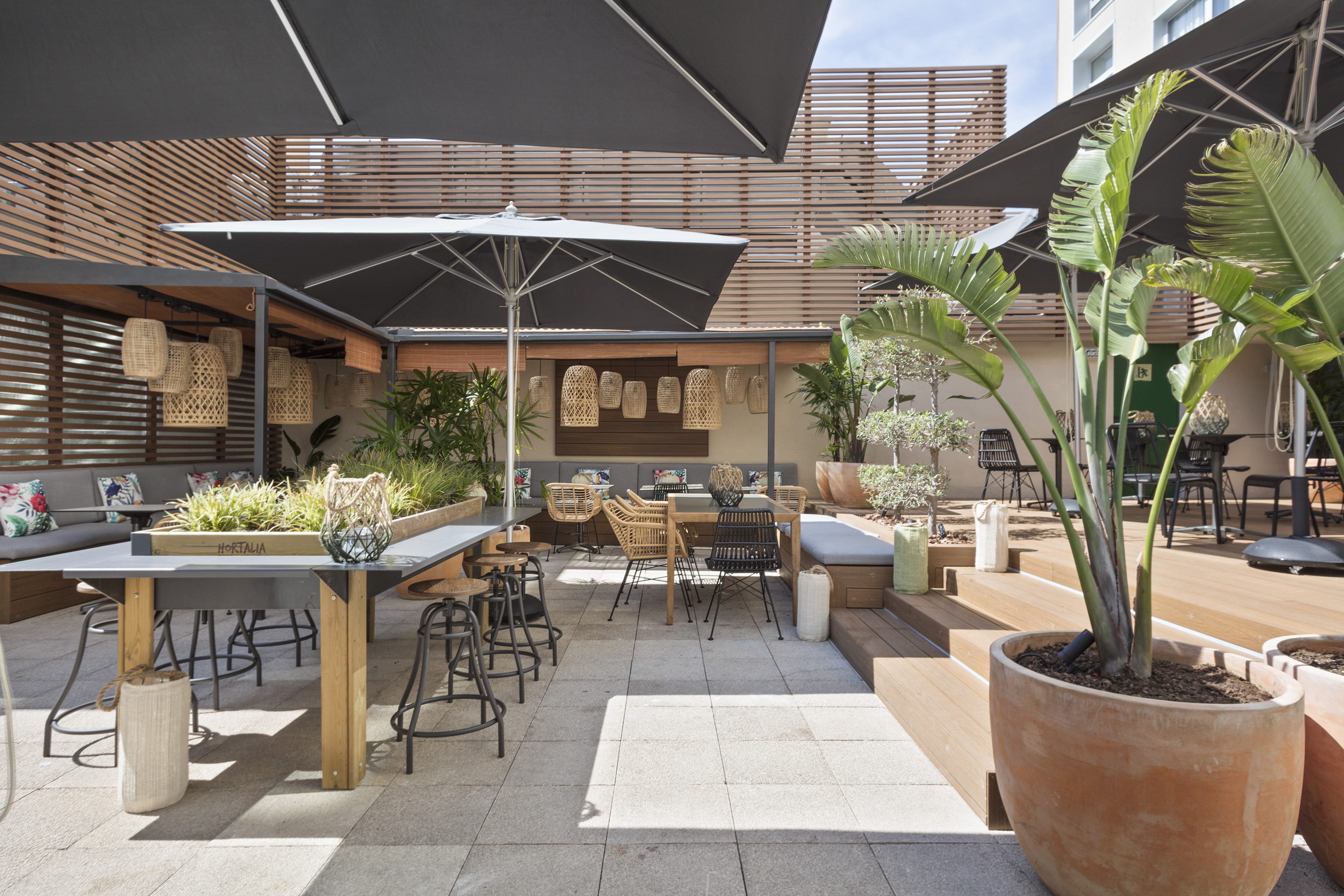 Outdoor patio dining area with shaded seating, tropical plants, and rattan lanterns.
