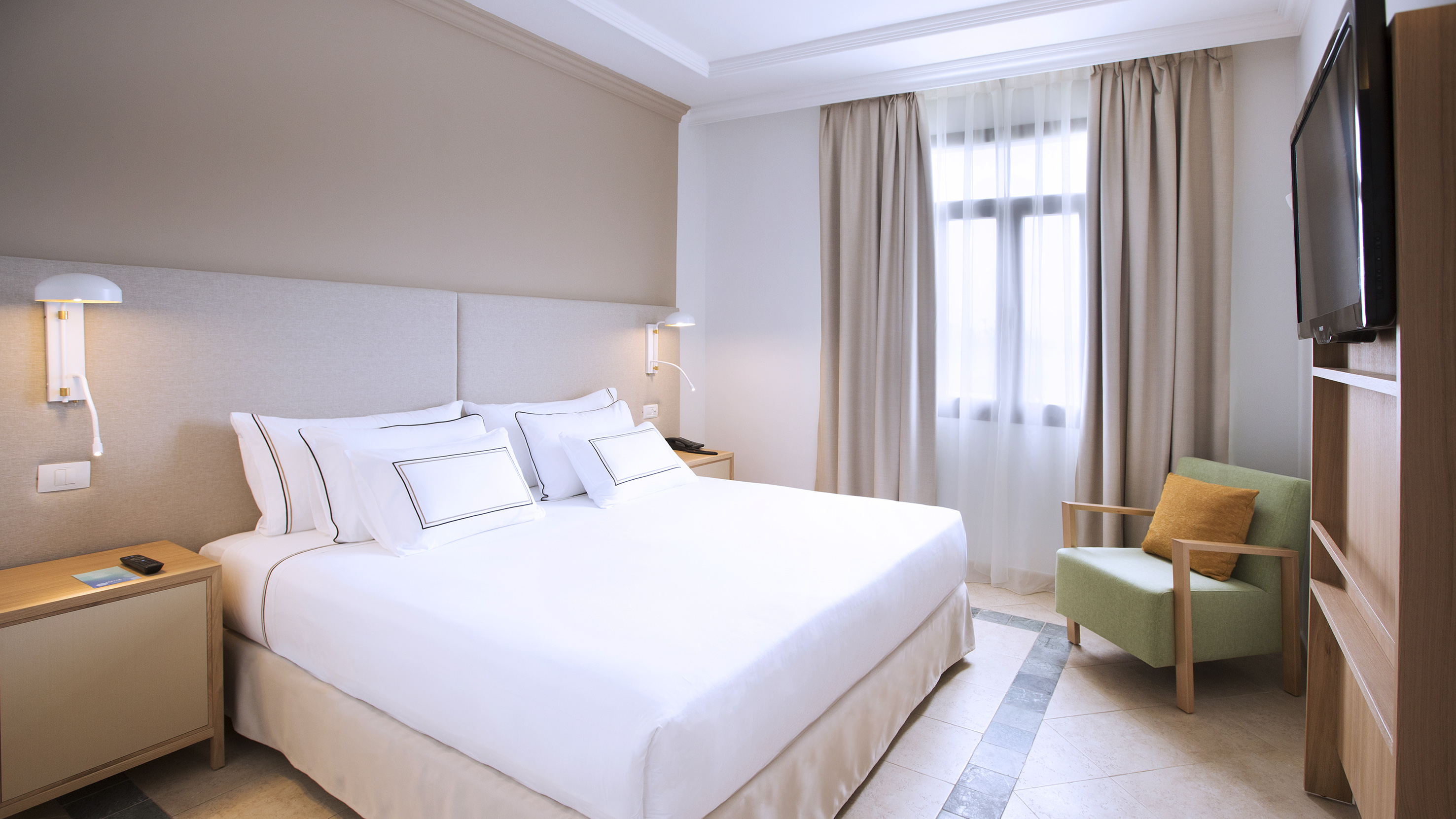 Modern hotel bedroom with white bed, sleek headboard, green armchair, and television.