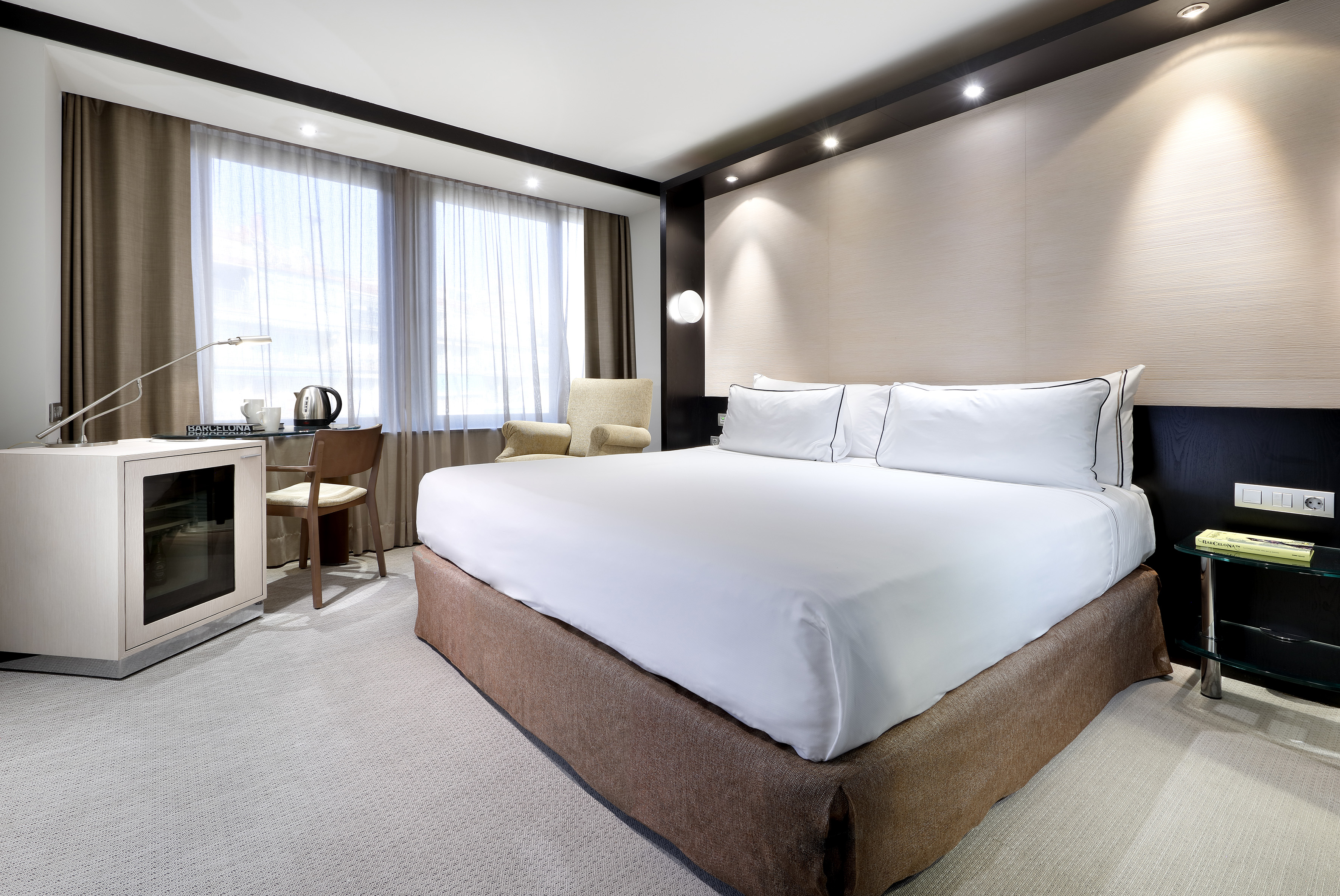 Modern hotel room with large bed, desk, mini-fridge, and bright windows.