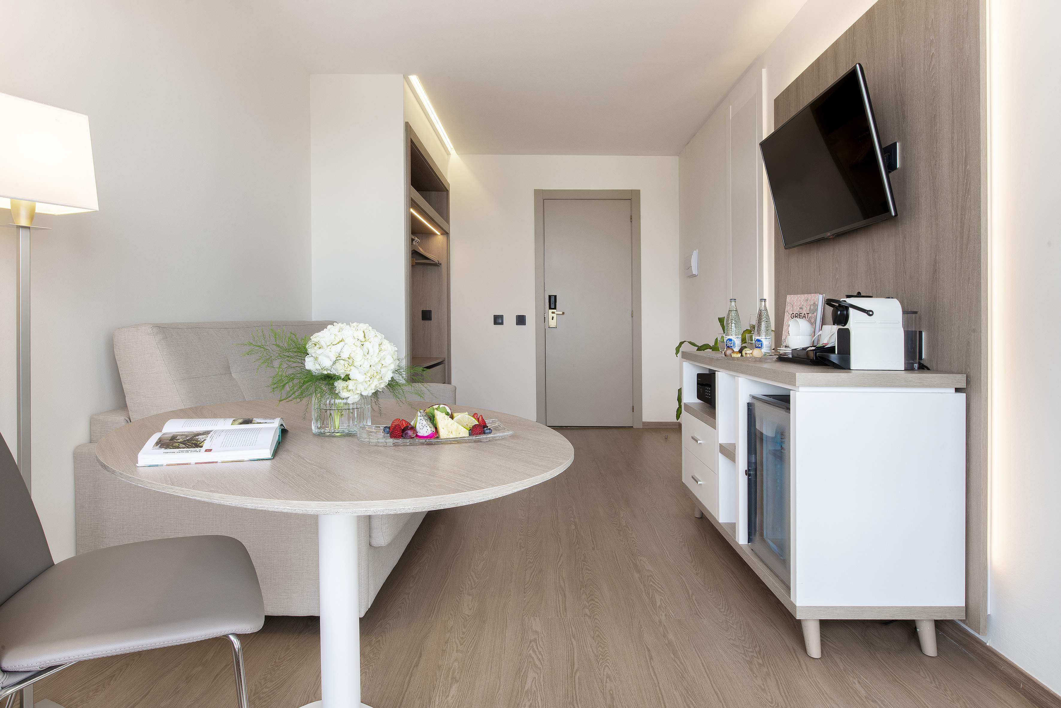 Modern hotel room with dining table, sofa, TV, Nespresso, and mini-bar.