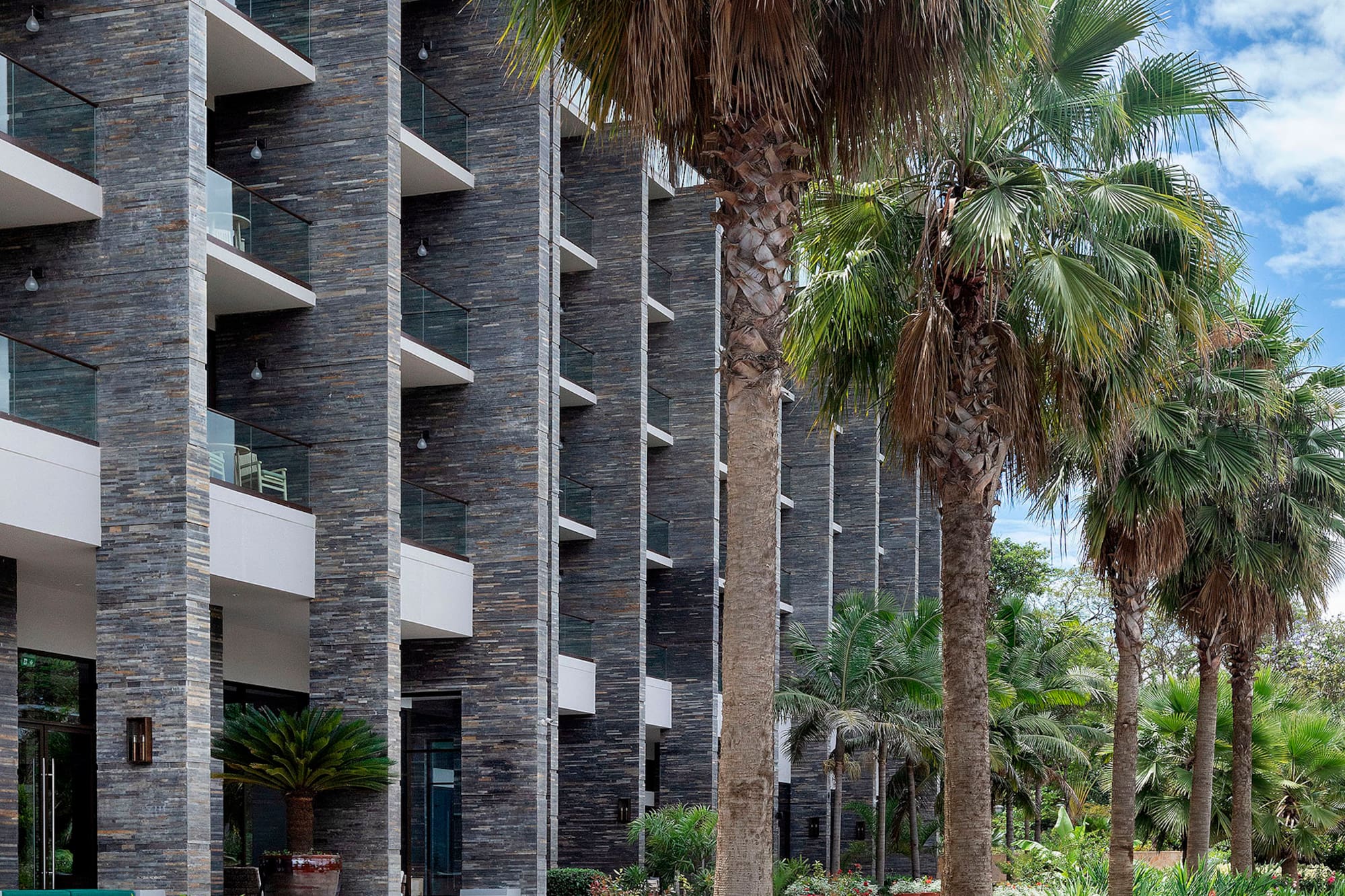 Modern resort building, stone facade, balconies, lush palm trees, patio.